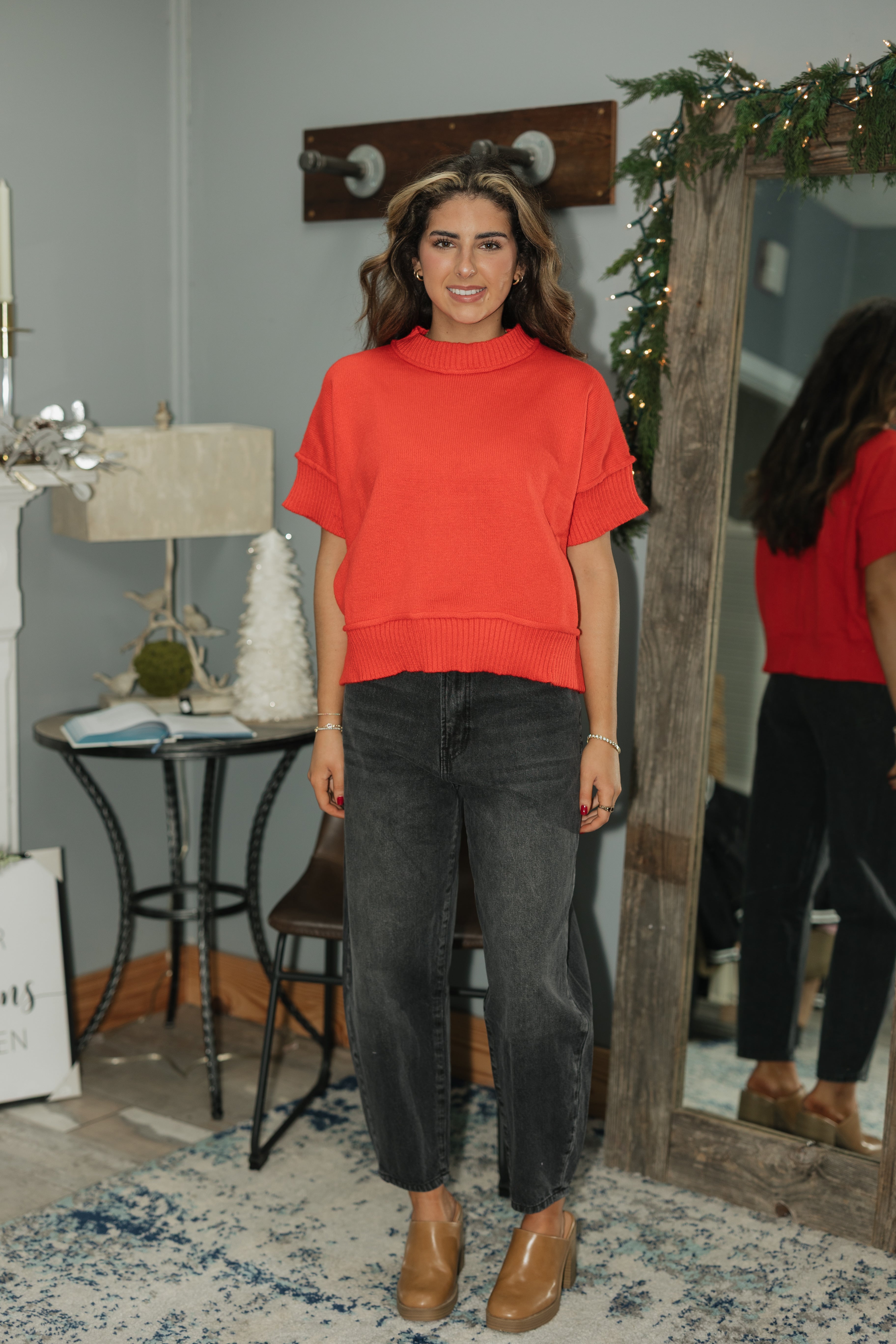Woman in a red top and black pants standing in a room with a mirror and decorative items.