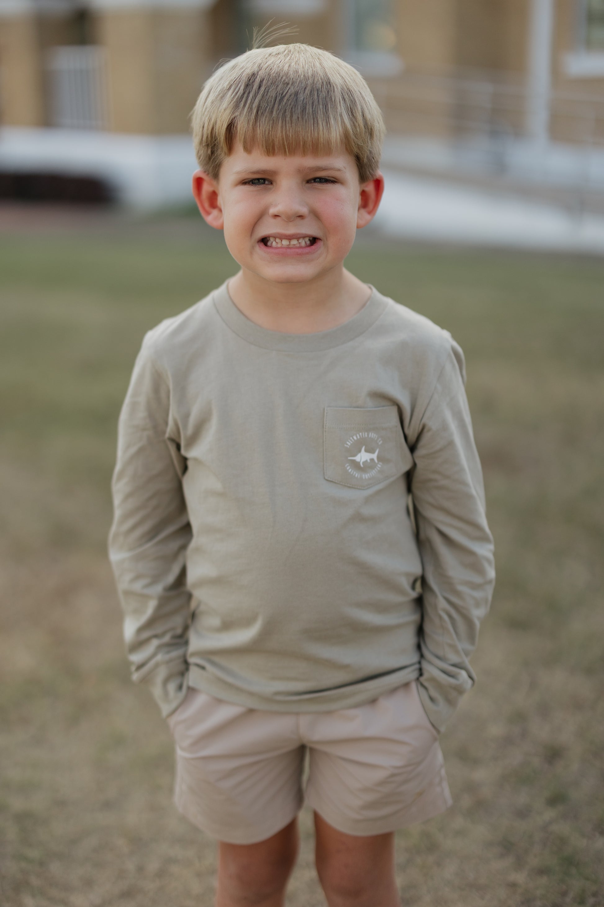 Young boy wearing a beige long-sleeve shirt and shorts outdoors.
