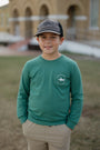 Young boy wearing a green long-sleeve shirt with a logo and a cap, standing outdoors.