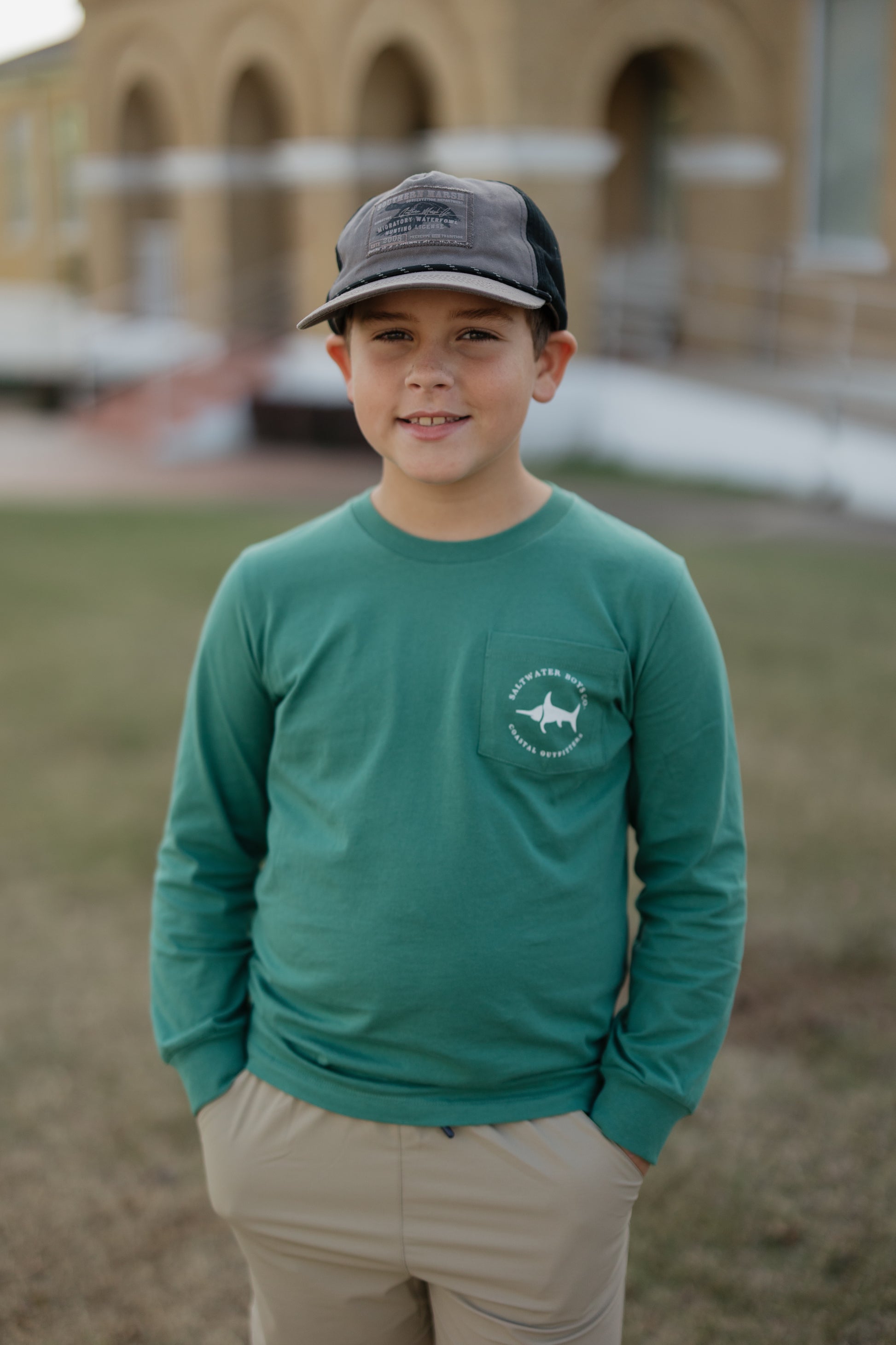 Young boy wearing a green long-sleeve shirt with a logo and a cap, standing outdoors.