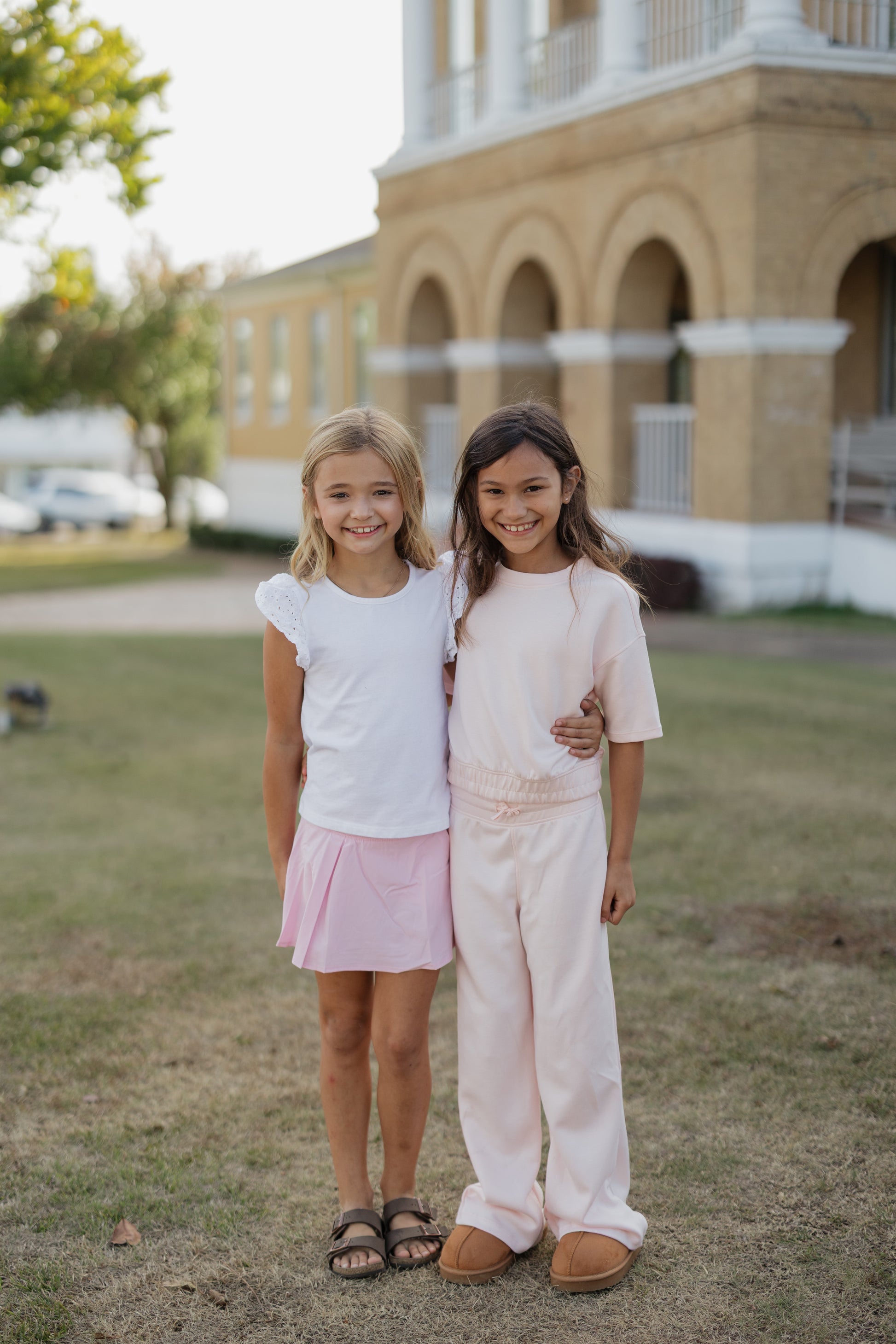 Two young girls standing together in a grassy area with a building in the background