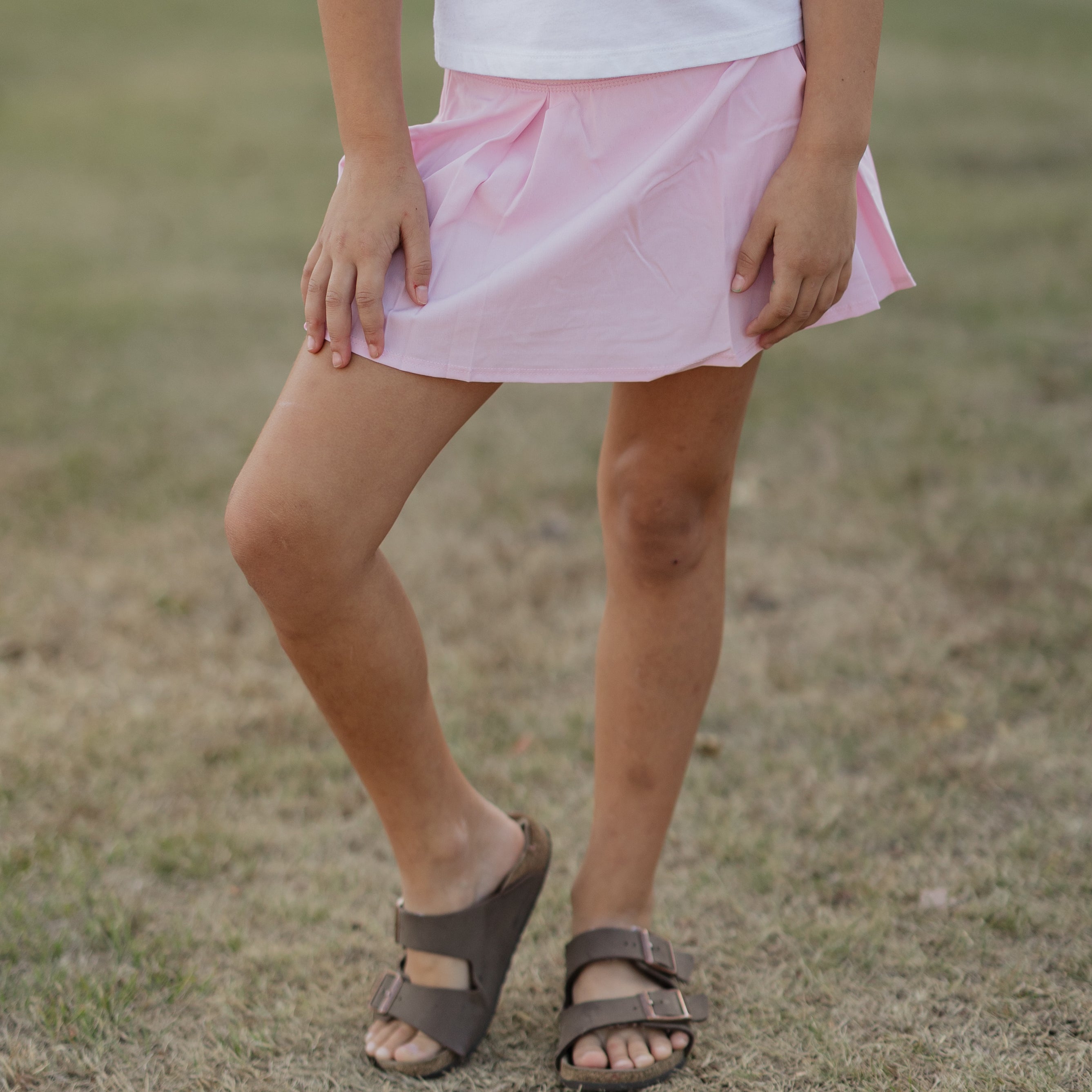 Person wearing a pink skirt and brown sandals on a grassy background