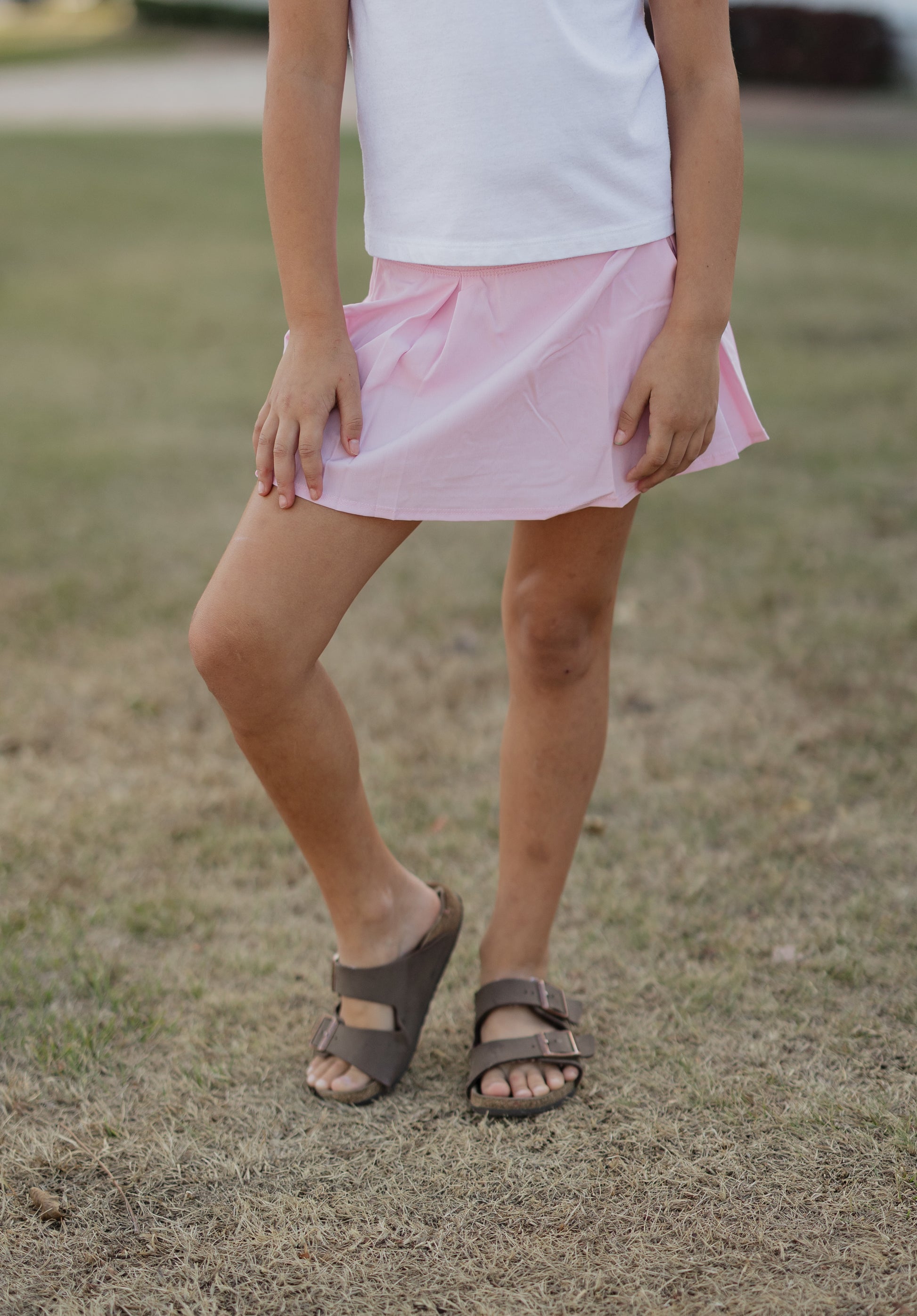 Person wearing a pink skirt and brown sandals on a grassy background
