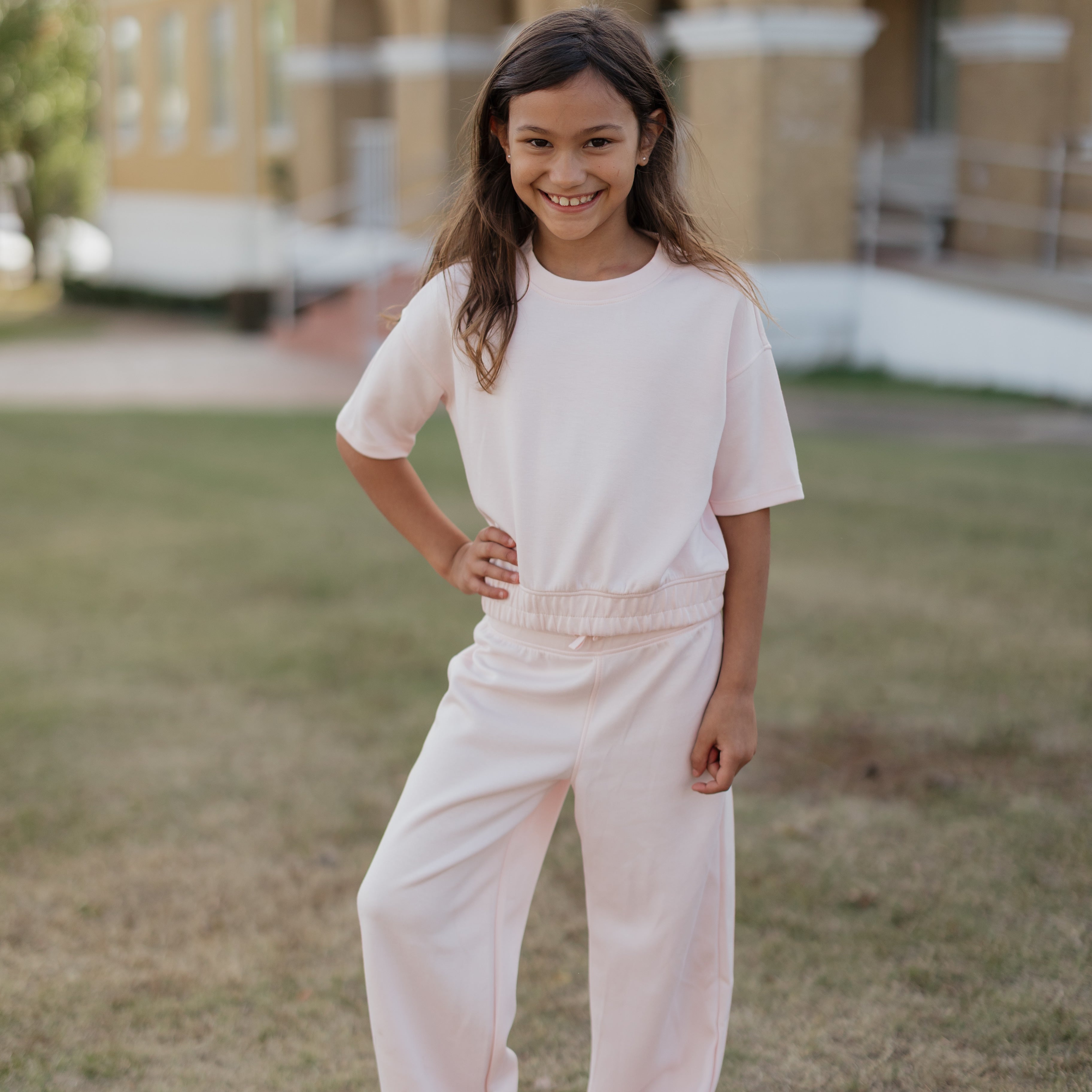 Young girl in a light pink outfit standing outdoors with a building in the background