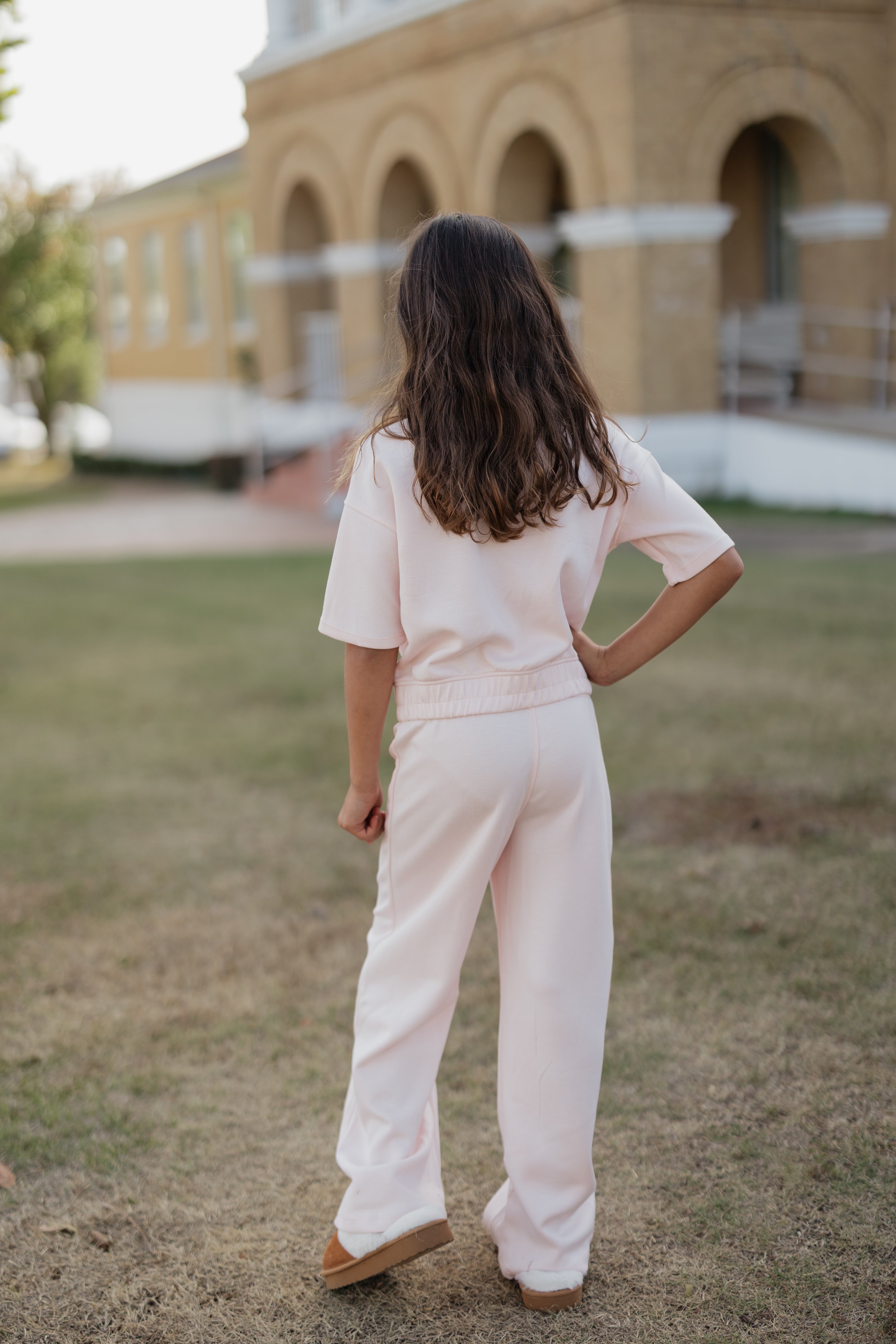 Person wearing a light pink outfit standing in a grassy area with a building in the background