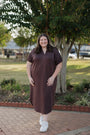 Woman wearing a brown dress standing outdoors on a brick path with trees in the background