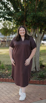 Woman wearing a brown dress standing outdoors on a brick path with trees in the background