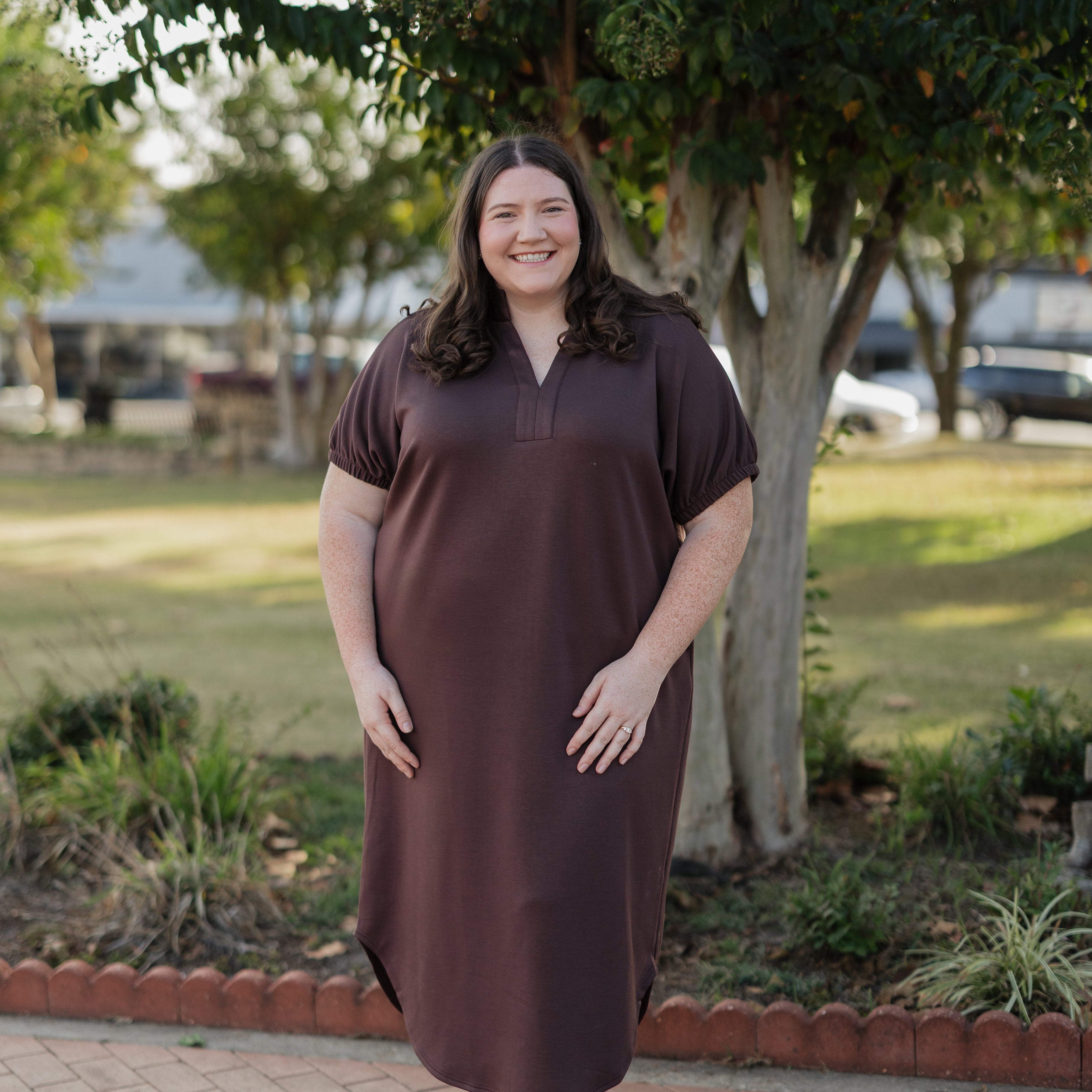 Woman wearing a brown dress standing outdoors on a brick path with trees in the background