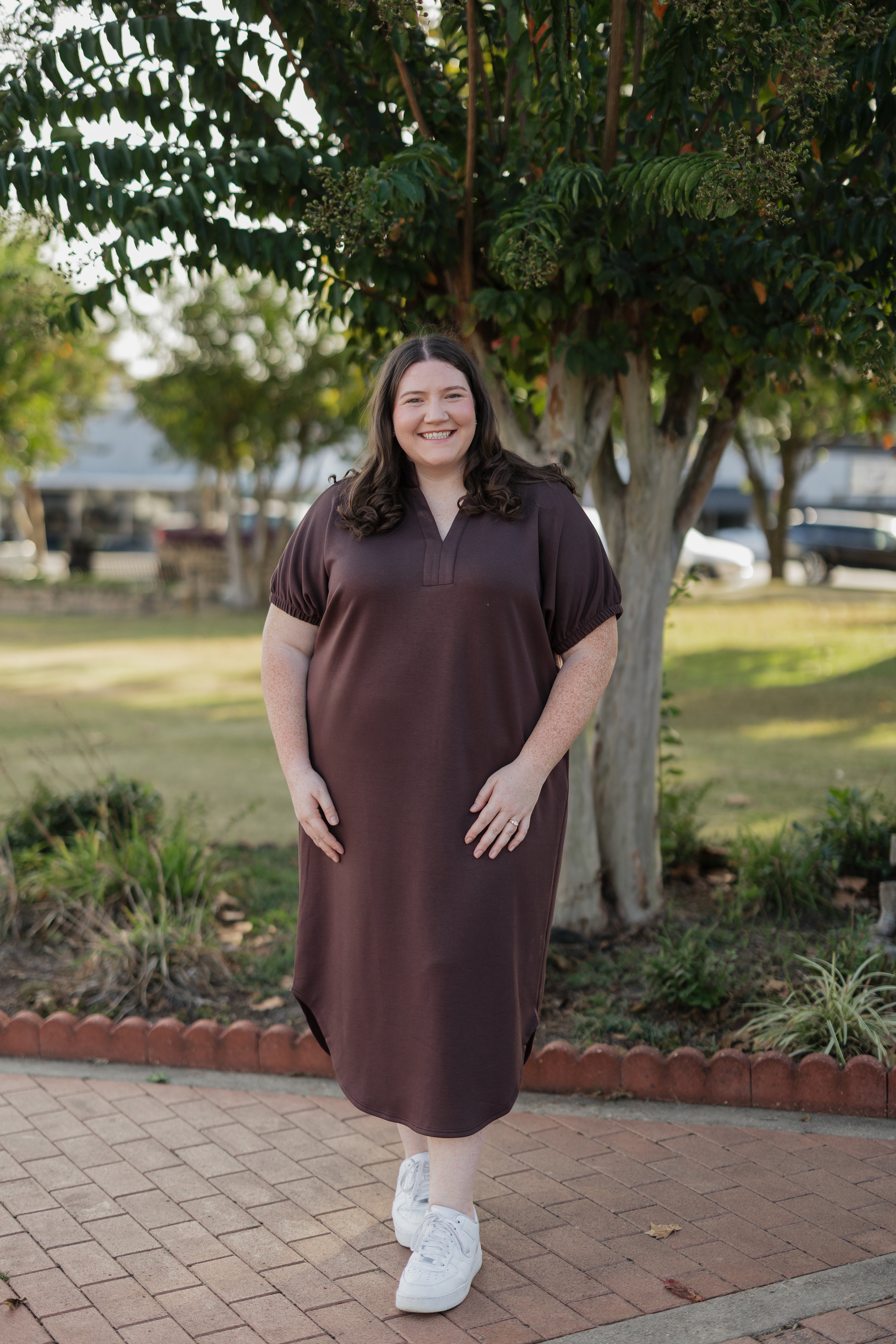 Woman wearing a brown dress standing outdoors on a brick path with trees in the background