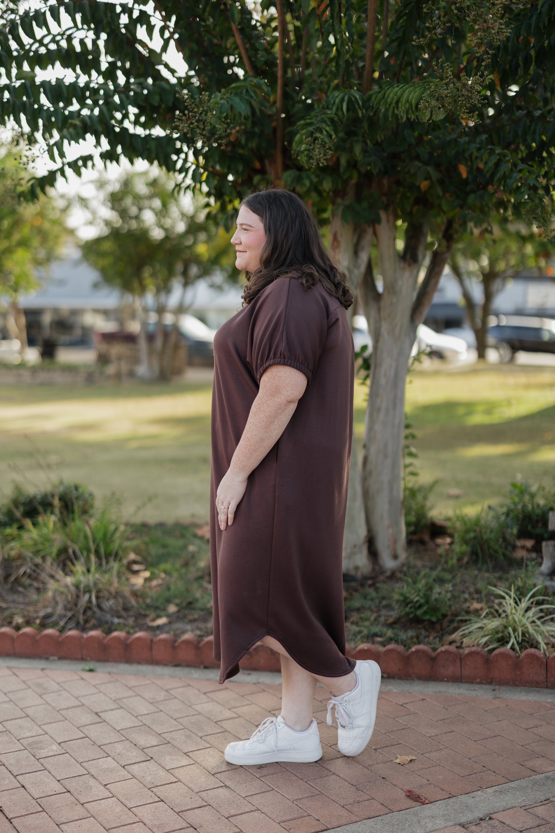 Woman in a brown dress standing on a sidewalk with trees and grass in the background