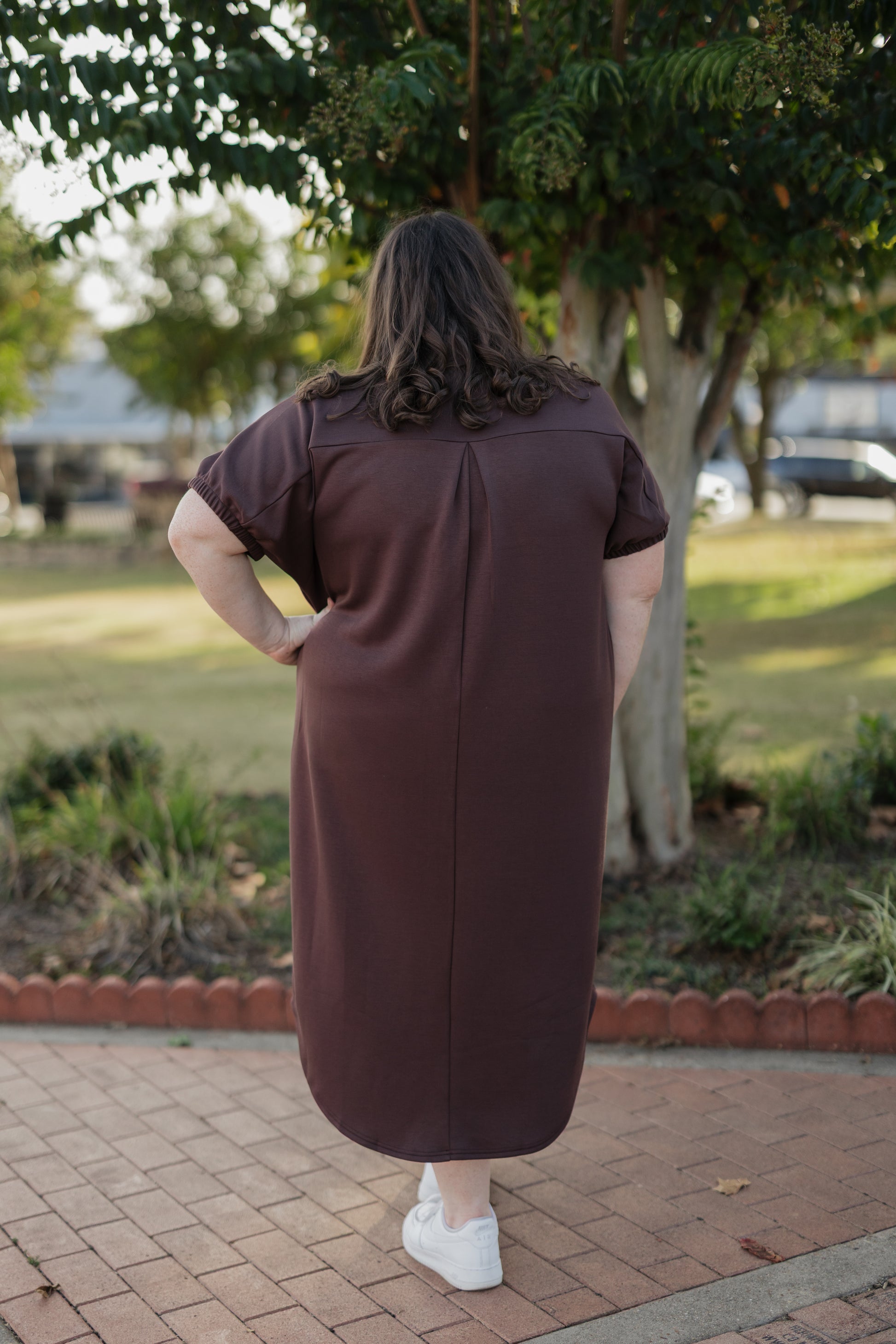 Person wearing a brown dress standing outdoors on a brick path with trees in the background