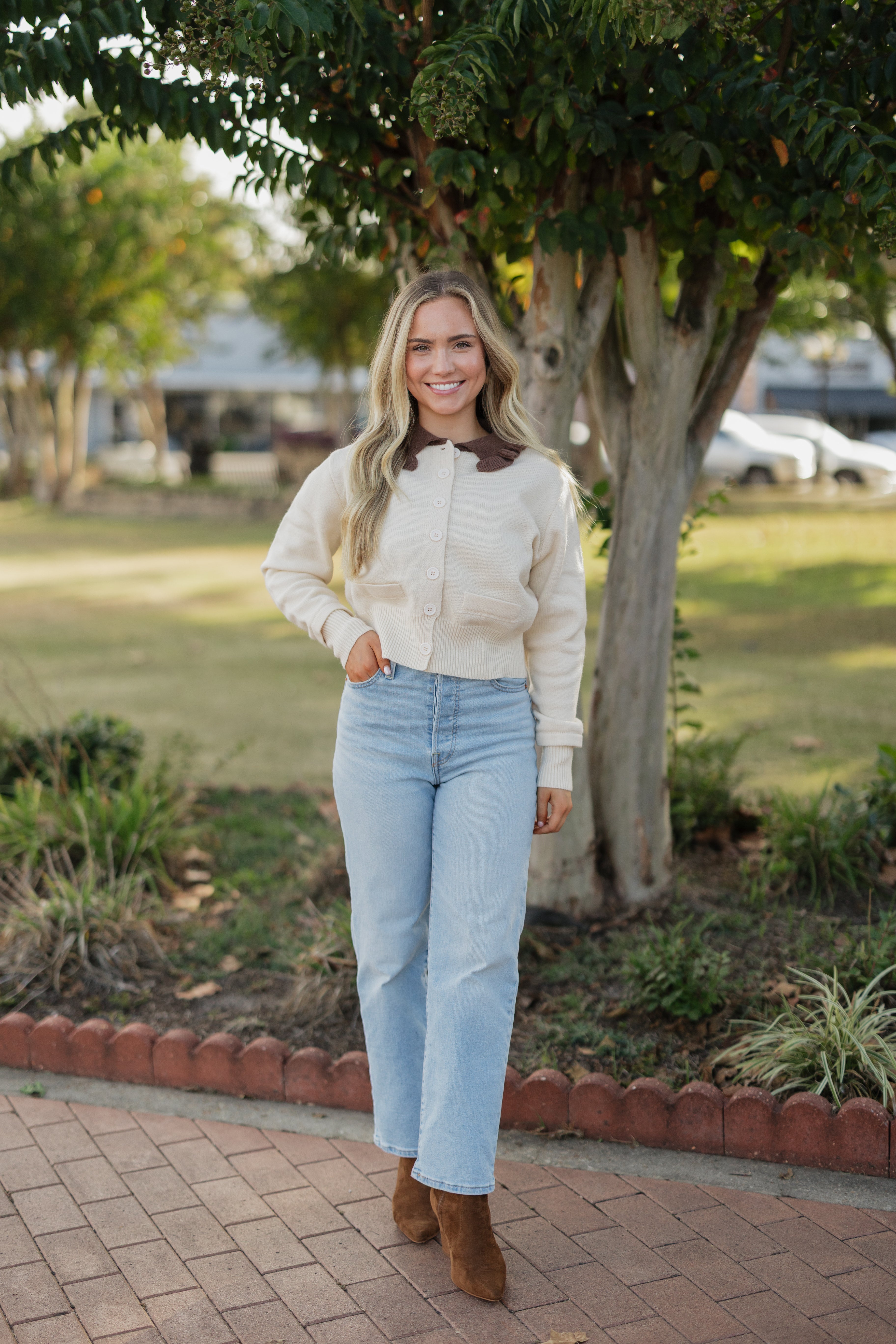 Woman in a beige jacket and light blue jeans standing outdoors near a tree