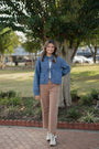 Woman in a blue jacket and beige pants standing outdoors near trees and a paved path.