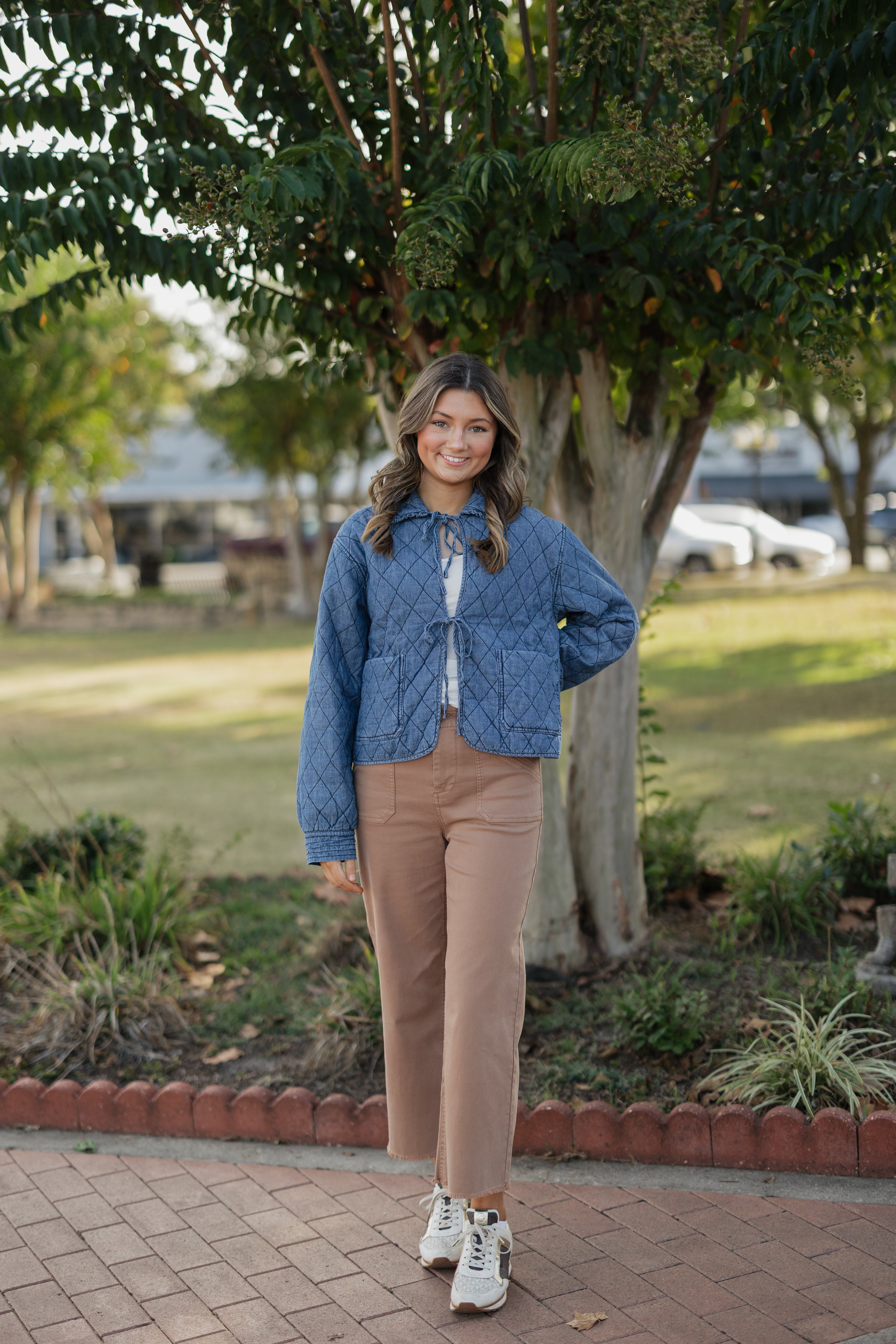 Woman in a blue jacket and beige pants standing outdoors near trees and a paved path.