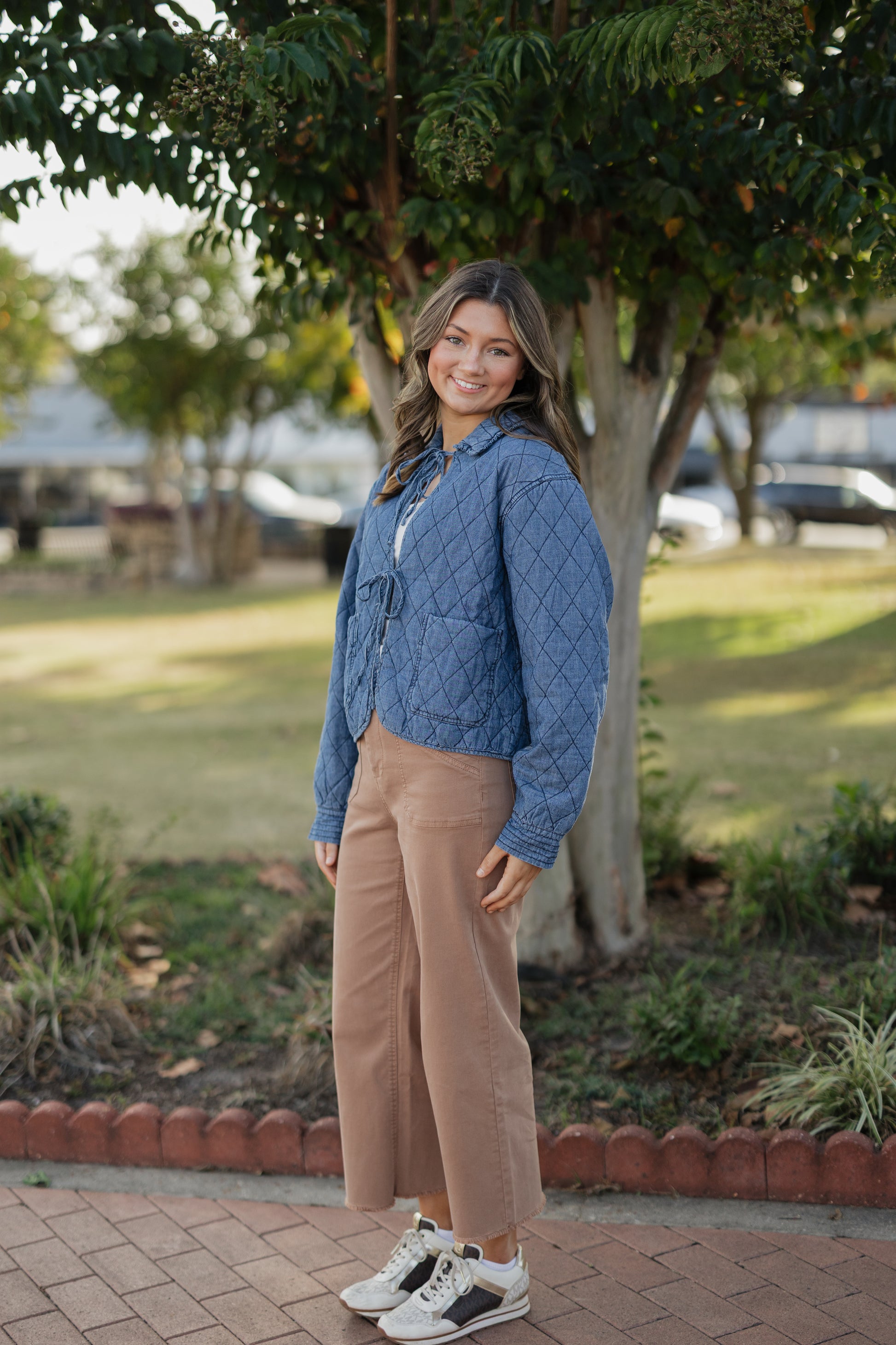 Woman in a blue jacket and beige pants standing outdoors on a brick path with trees in the background.