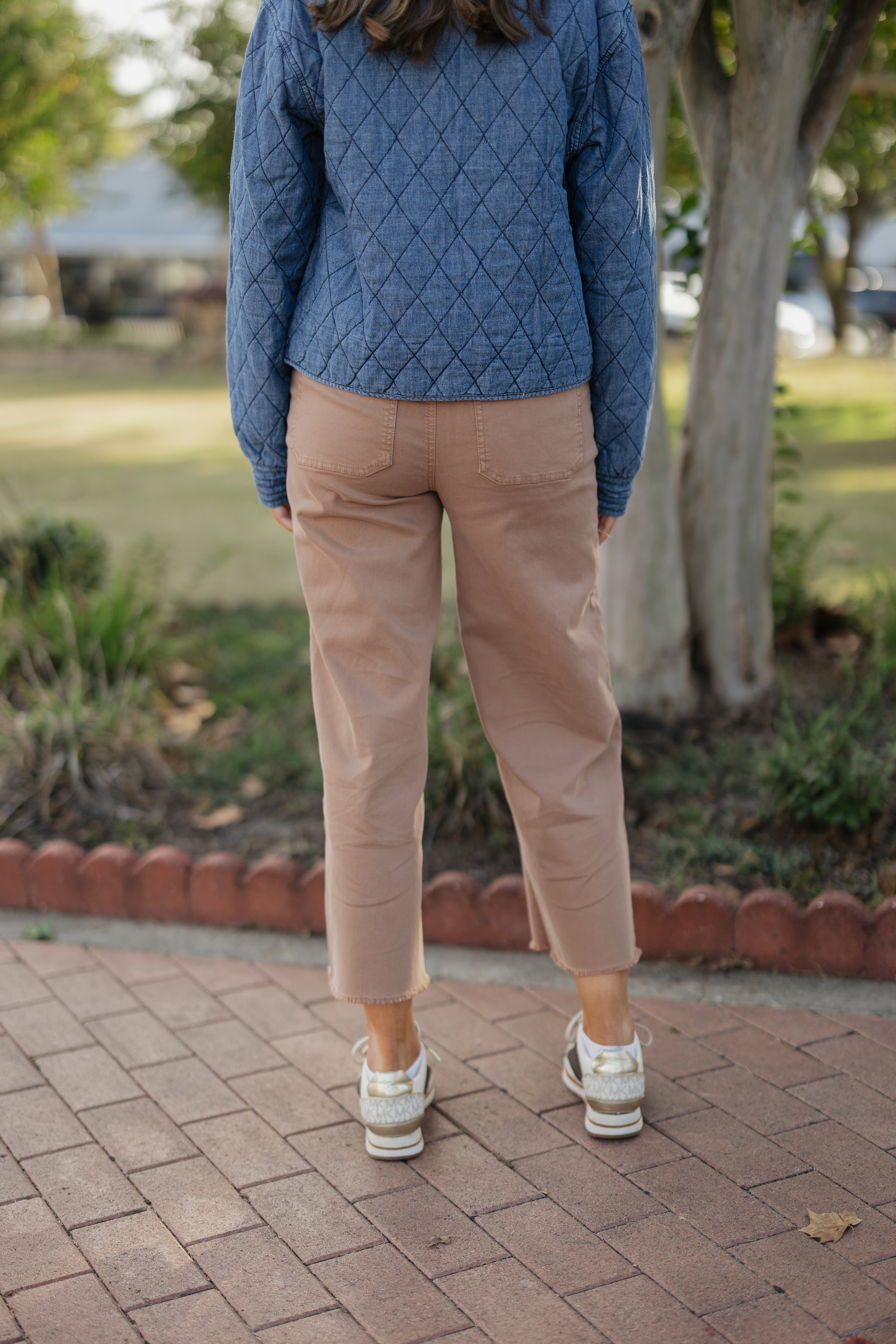 Person wearing a blue quilted jacket and beige pants standing on a brick path with a blurred background