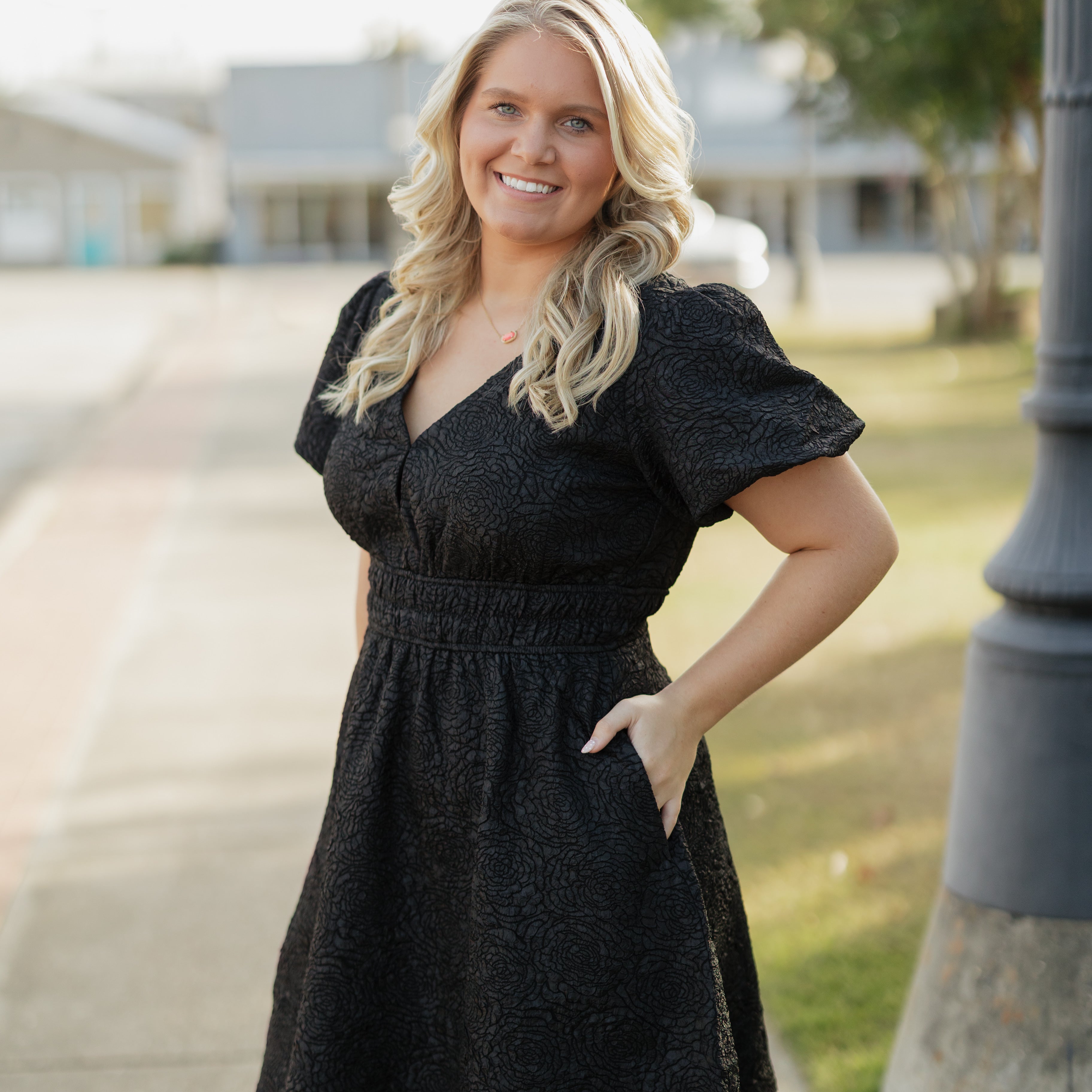 Woman in a black dress standing outdoors on a sidewalk with grass and trees in the background.