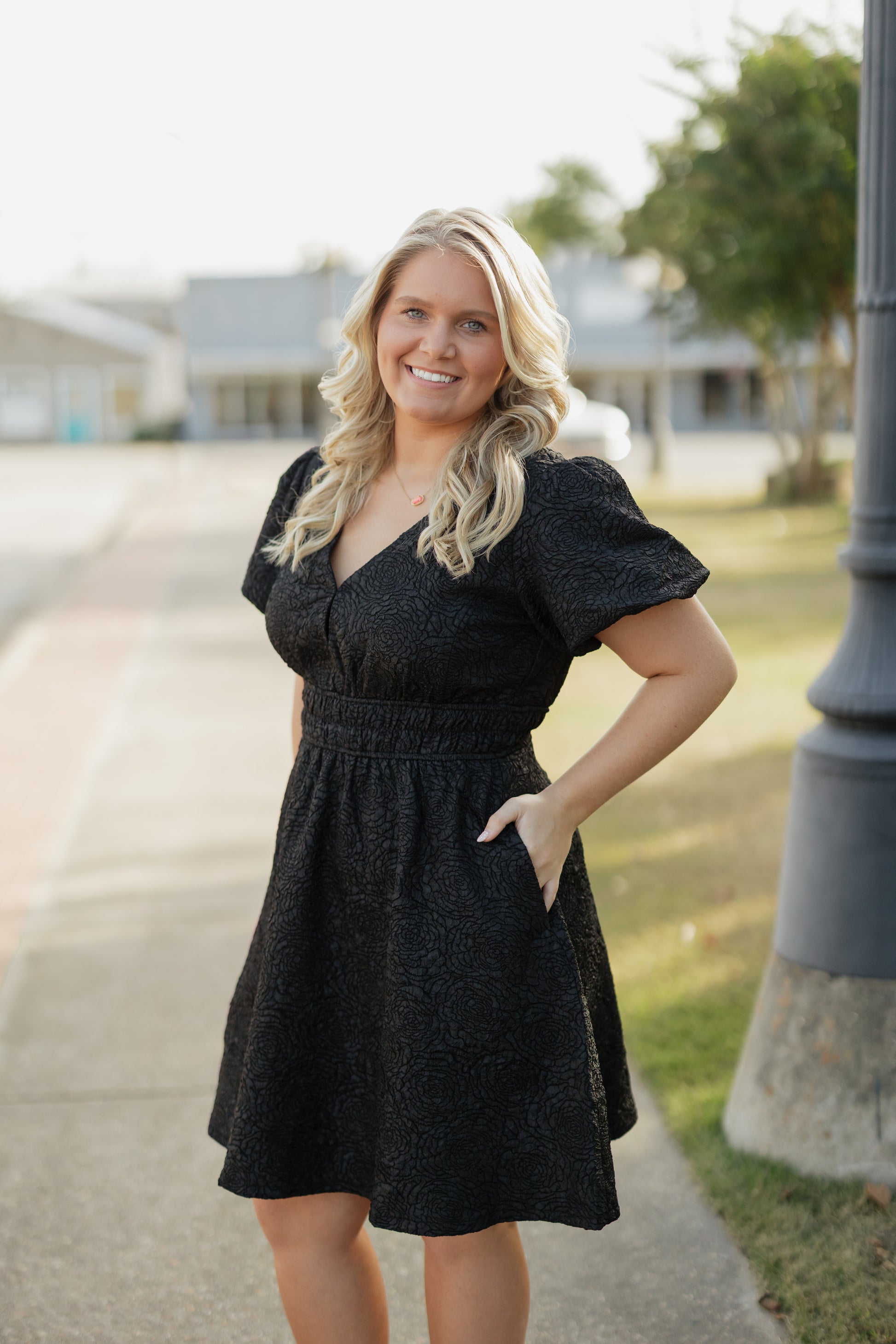 Woman in a black dress standing outdoors on a sidewalk with grass and trees in the background.