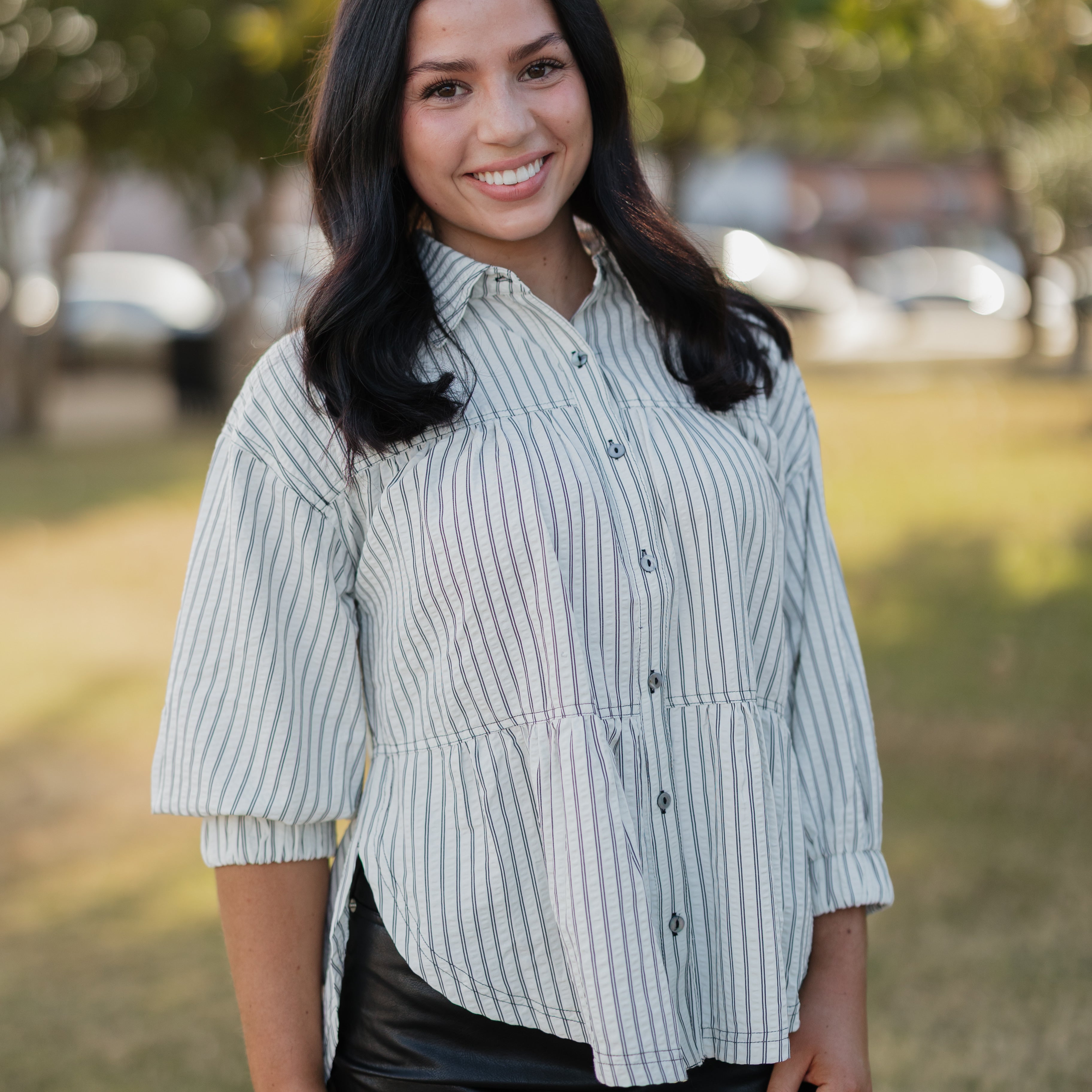 Woman wearing a striped shirt and black skirt standing outdoors with trees and cars in the background