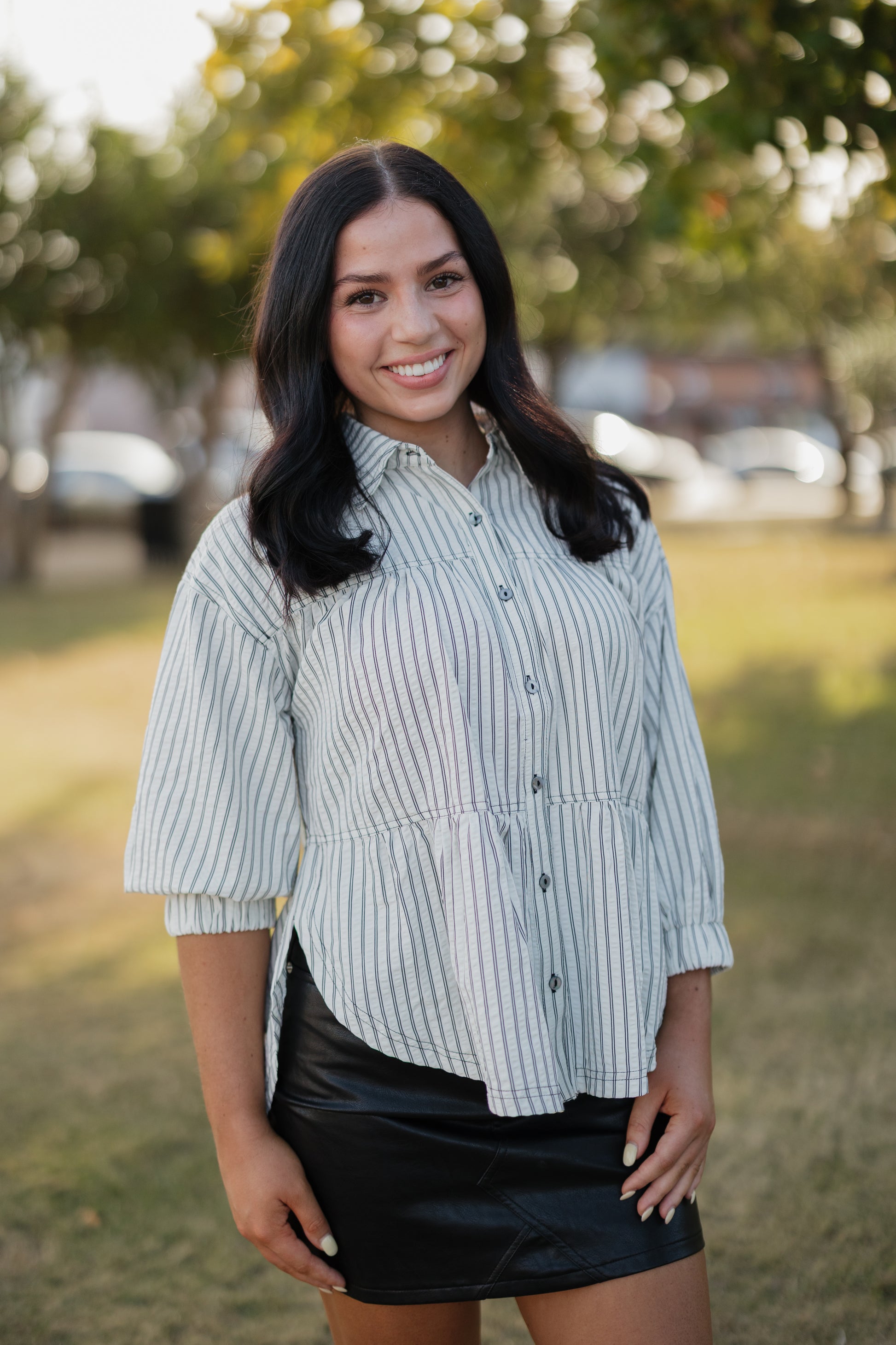 Woman wearing a striped shirt and black skirt standing outdoors with trees and cars in the background