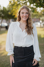 Woman wearing a white blouse and black pants standing outdoors with trees in the background