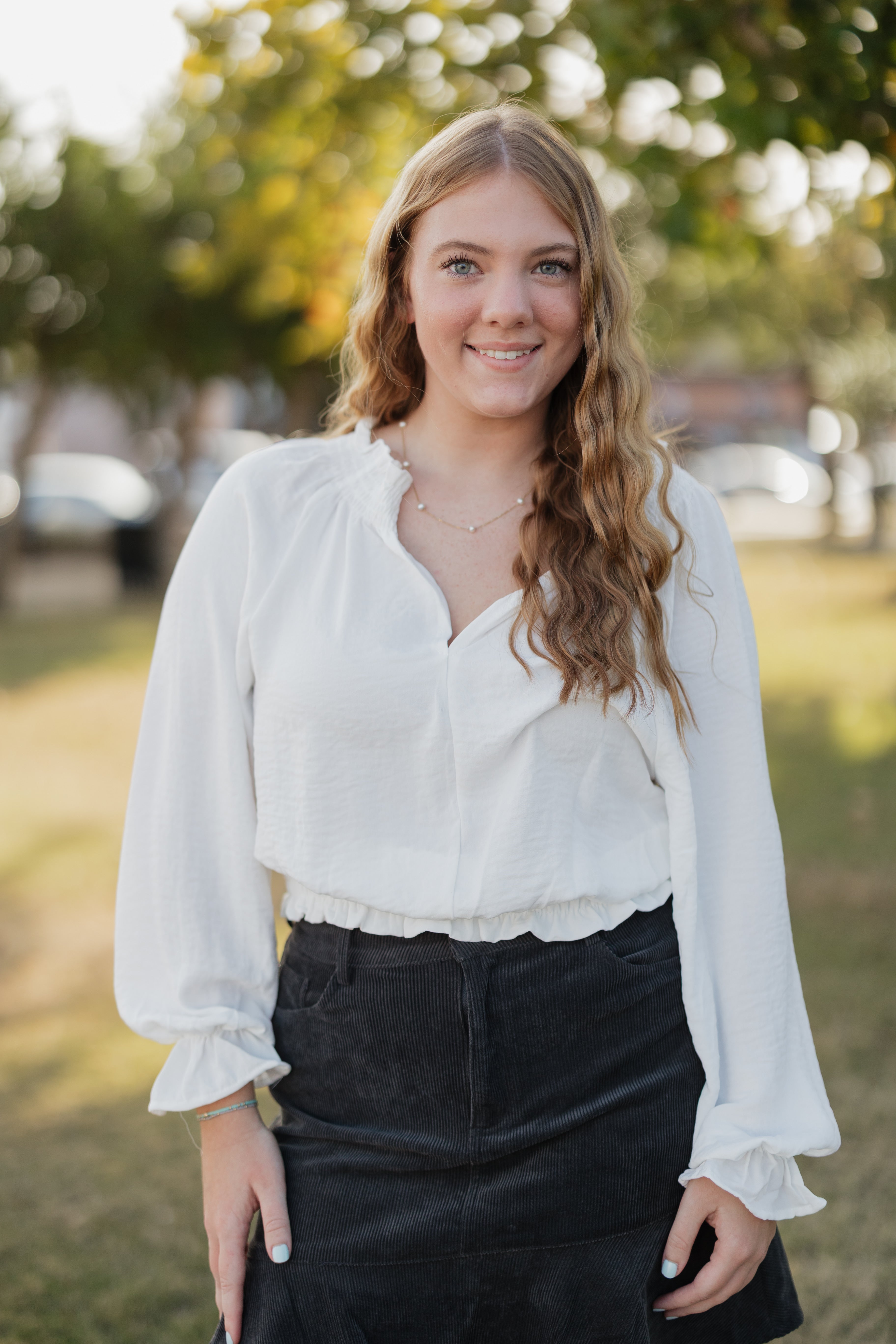 Woman wearing a white blouse and black pants standing outdoors with trees in the background
