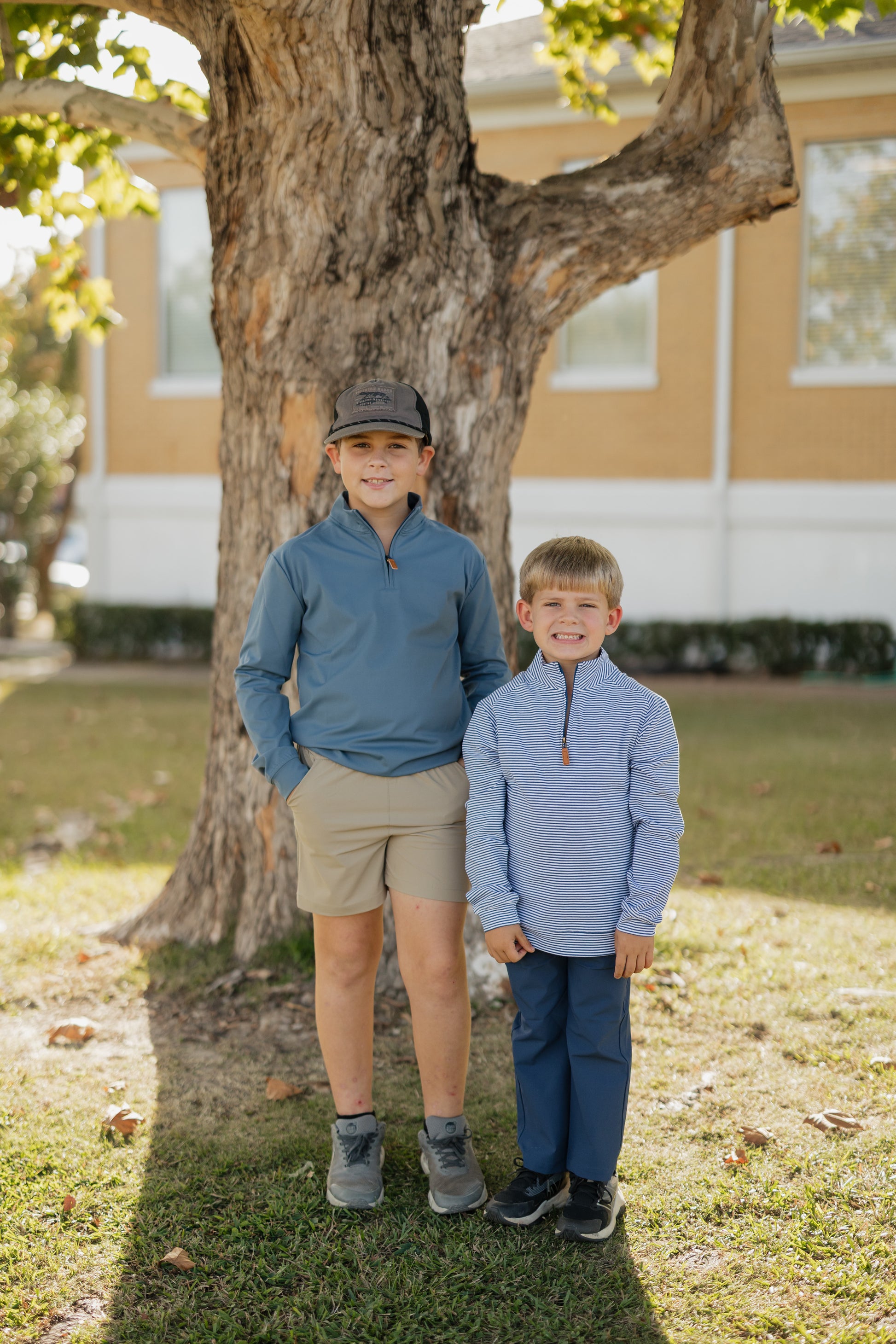 Two boys standing under a tree in a grassy area with a building in the background.