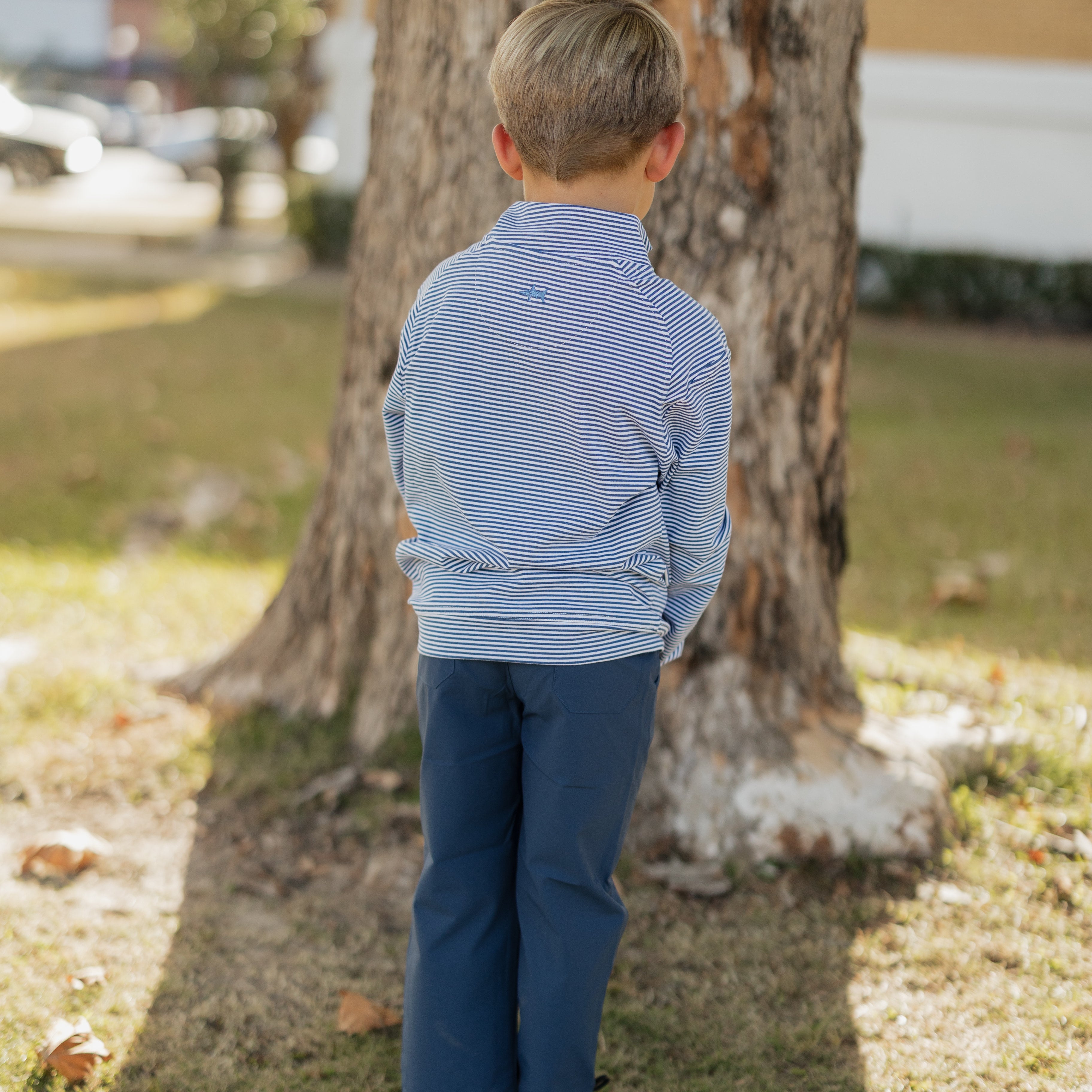 Child in a striped shirt and blue pants standing next to a tree