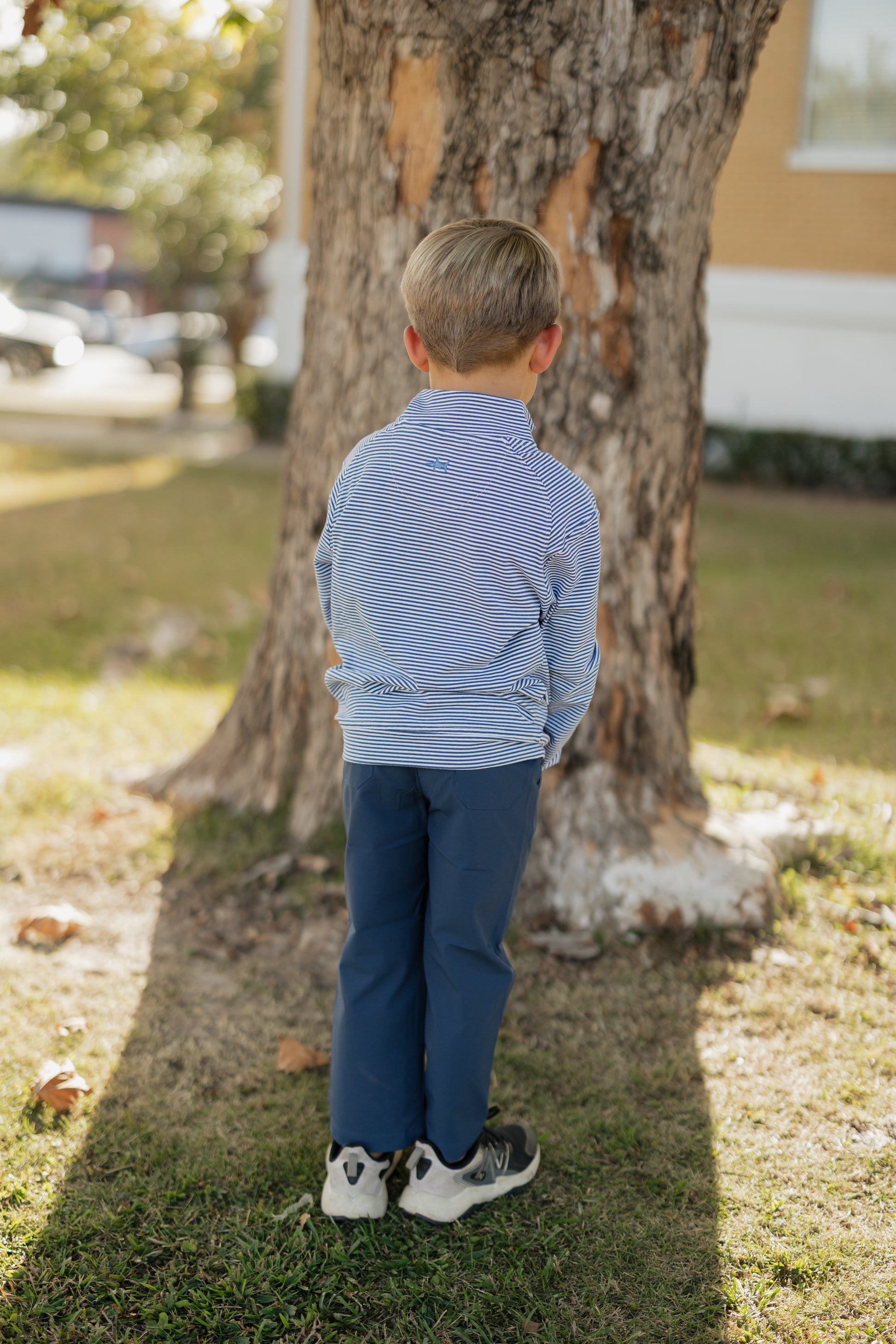 Child in a striped shirt and blue pants standing next to a tree