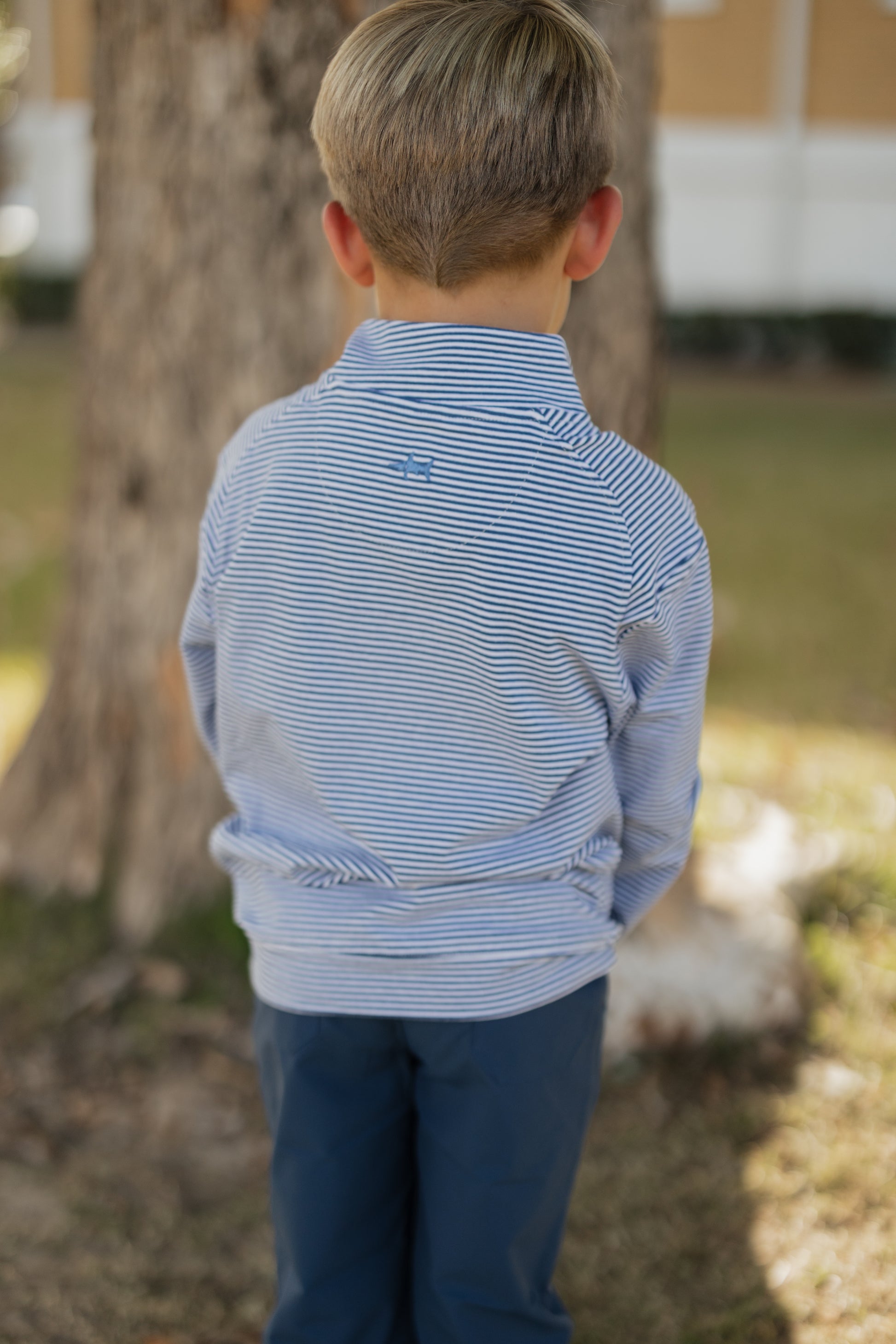 Child wearing a blue striped shirt and dark pants standing outdoors with trees and grass in the background