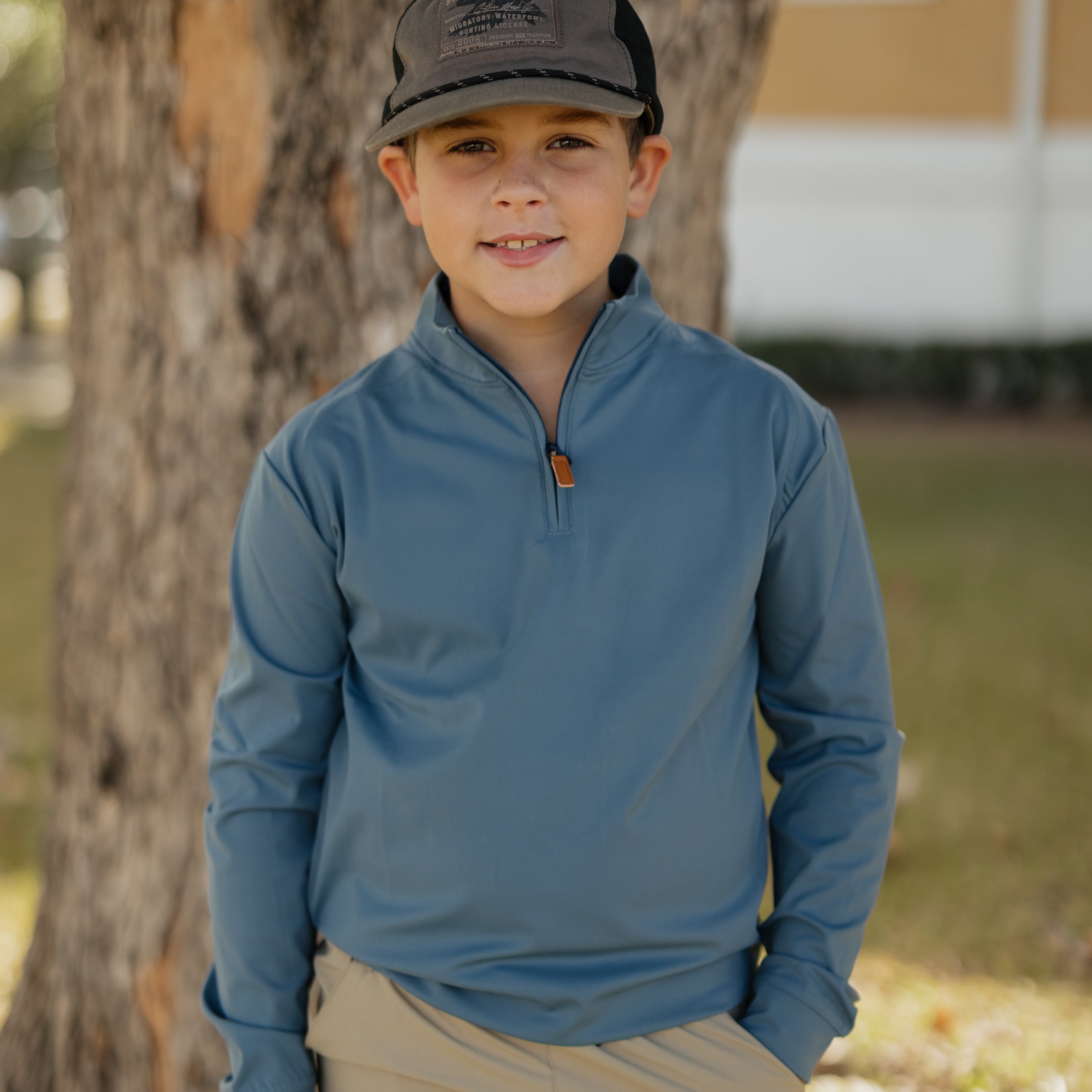 Child wearing a blue long-sleeve shirt and beige shorts standing next to a tree.