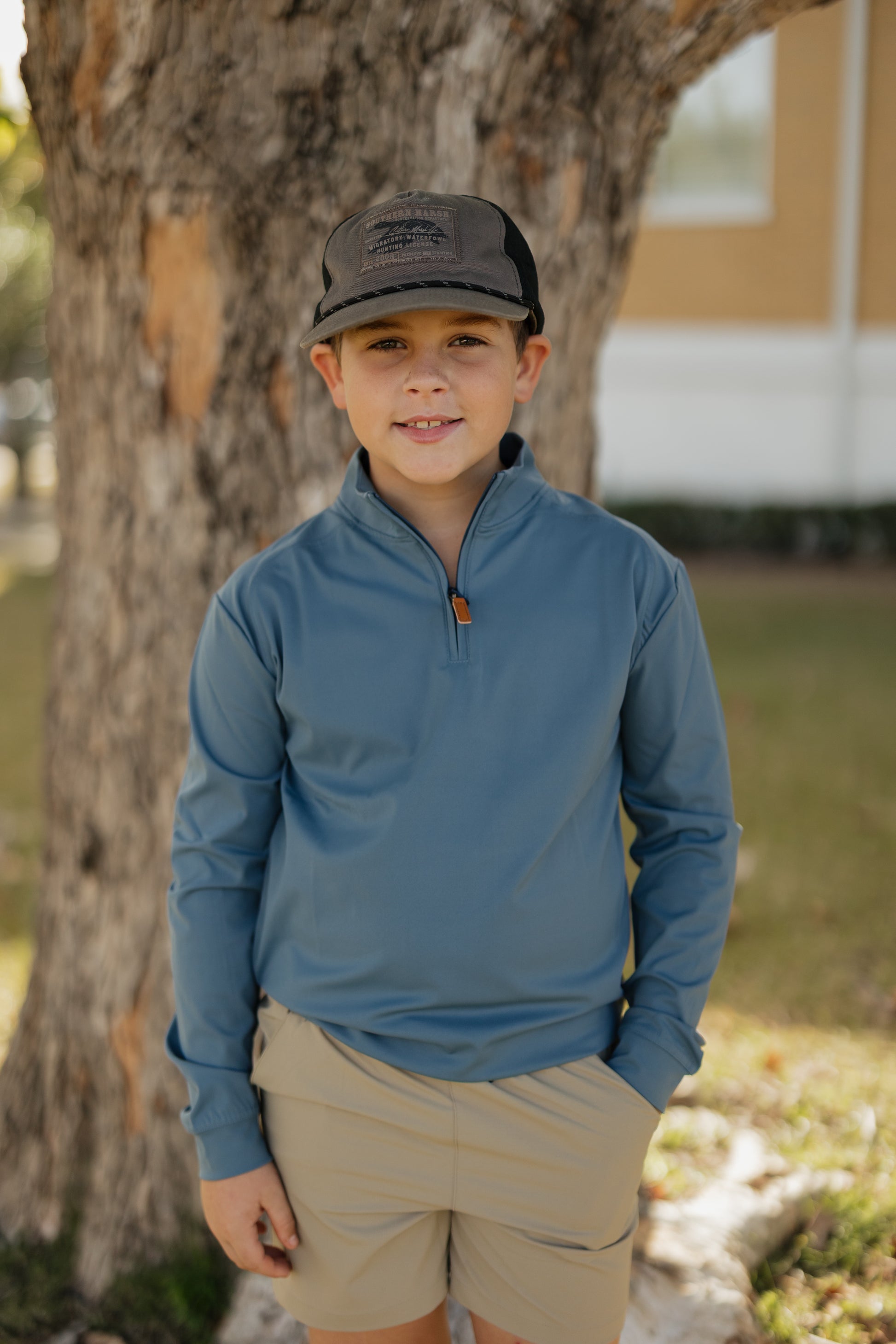 Child wearing a blue long-sleeve shirt and beige shorts standing next to a tree.