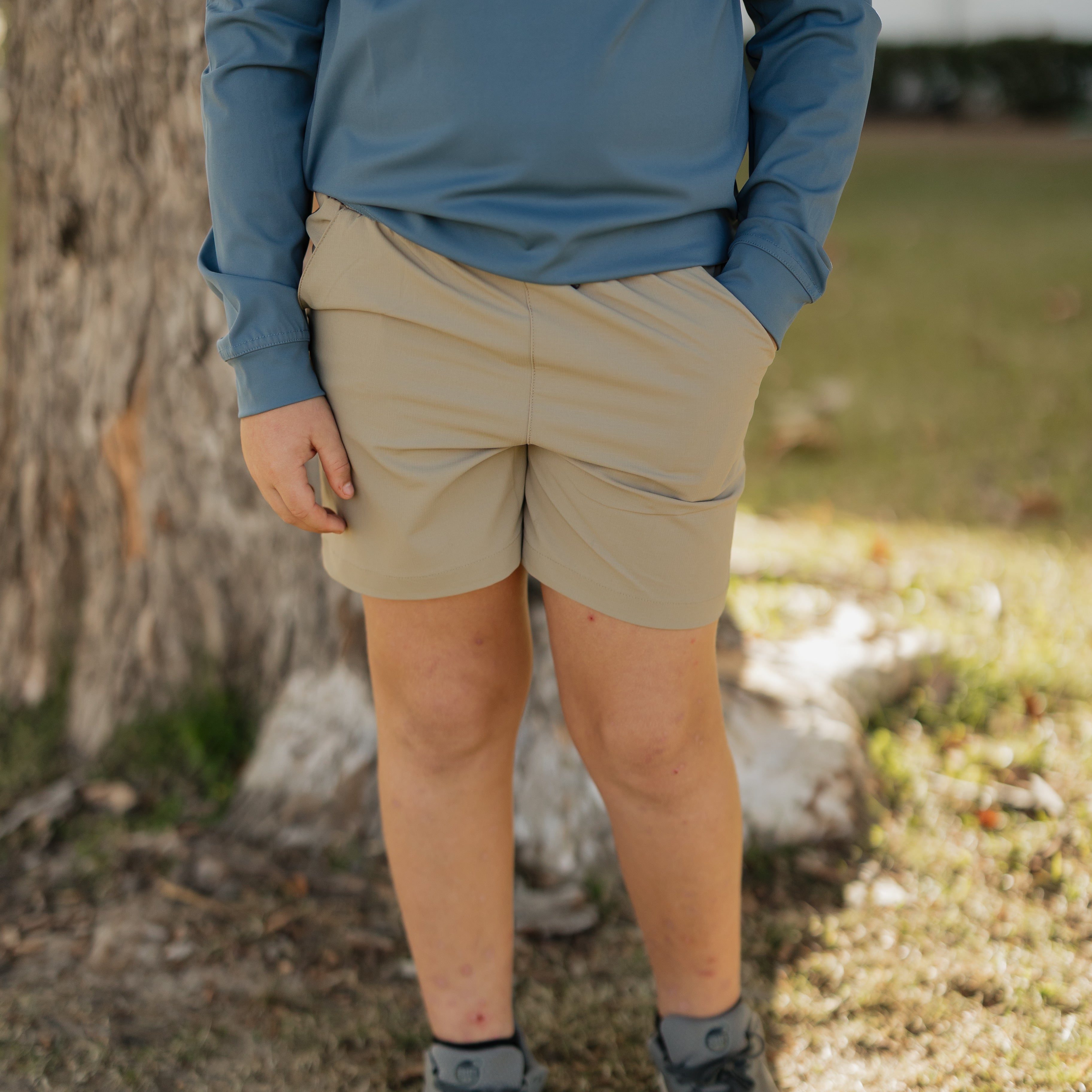 Person wearing a blue long-sleeve shirt, beige shorts, and gray shoes standing outdoors near a tree.