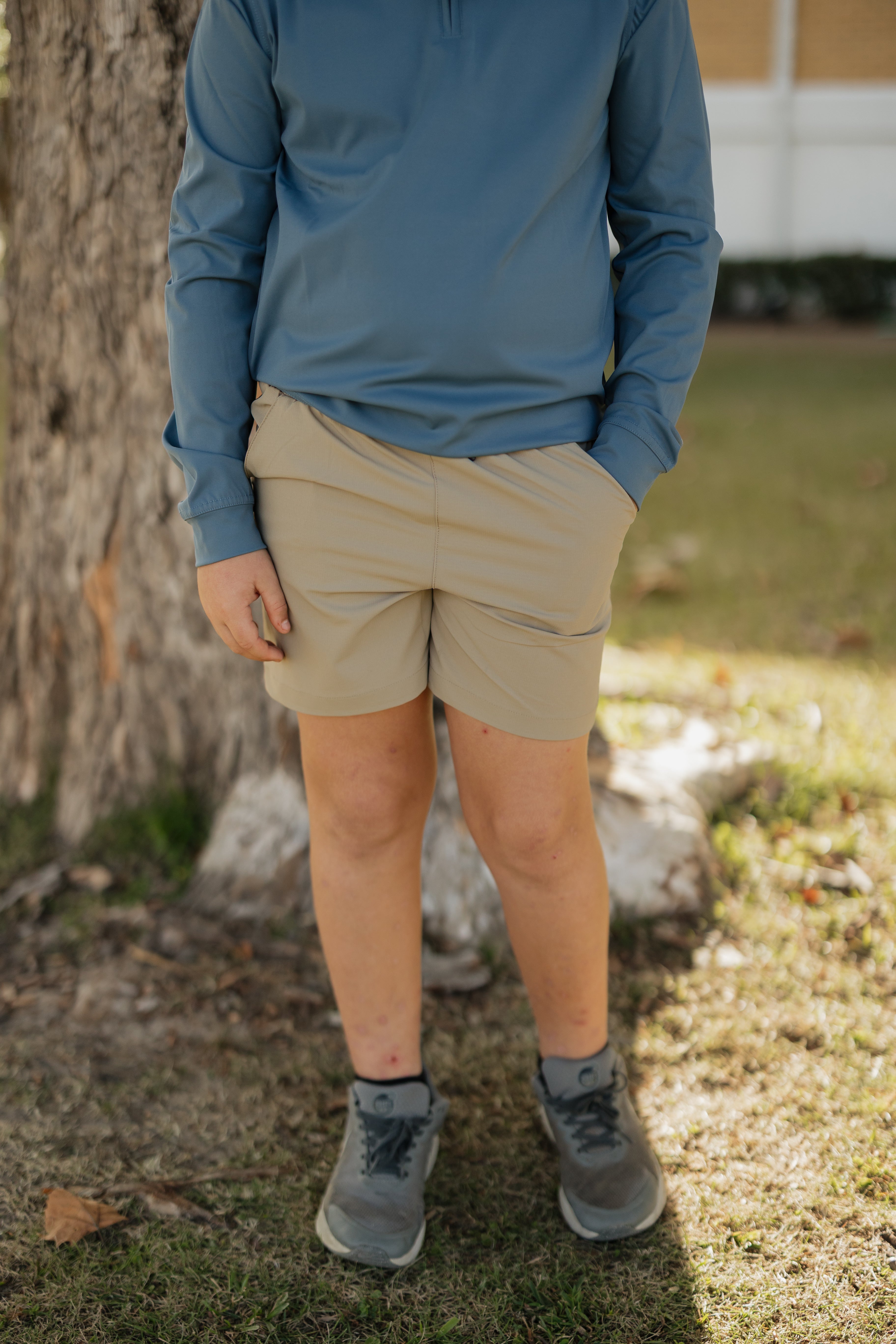 Person wearing a blue long-sleeve shirt, beige shorts, and gray shoes standing outdoors near a tree.