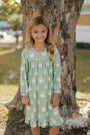 Young girl wearing a green dress with Santa Claus pattern standing next to a tree.