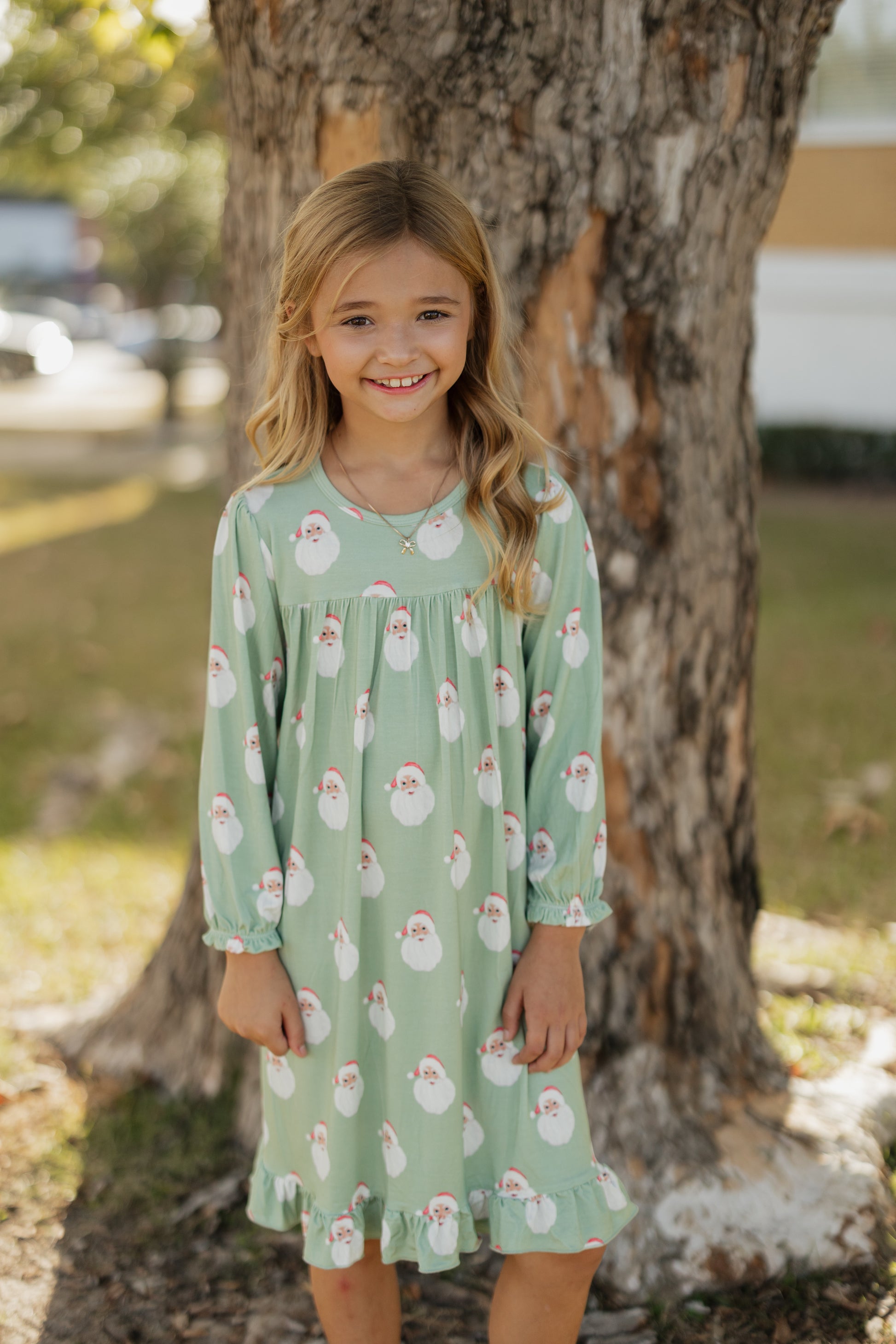 Young girl wearing a green dress with Santa Claus pattern standing next to a tree.