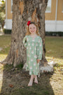 Young girl in a green dress with a red bow standing next to a tree