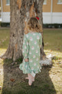 Child wearing a green dress with Santa Claus pattern standing near a tree