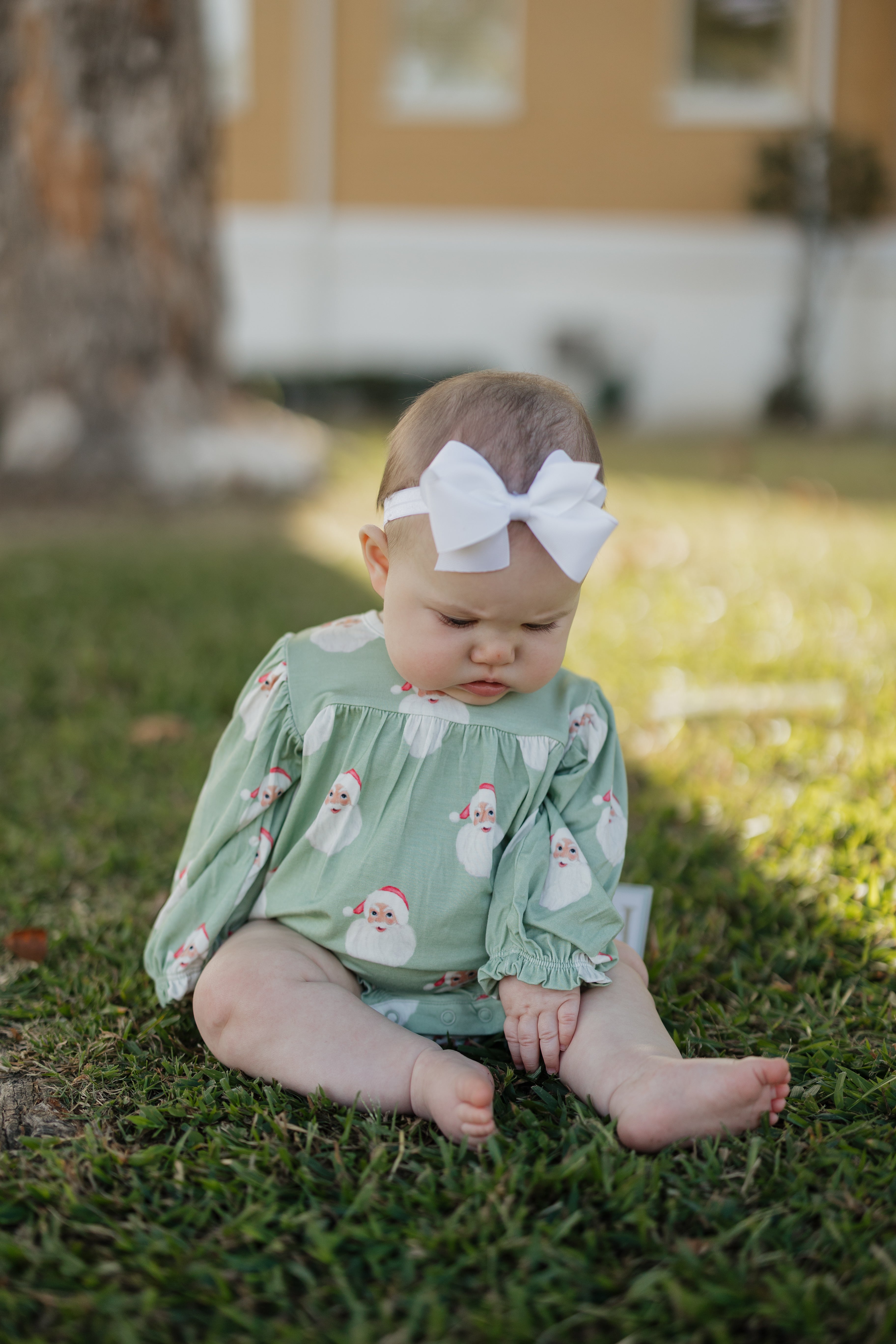 Baby sitting on grass wearing a green bubble romper with floral patterns and a white headband.