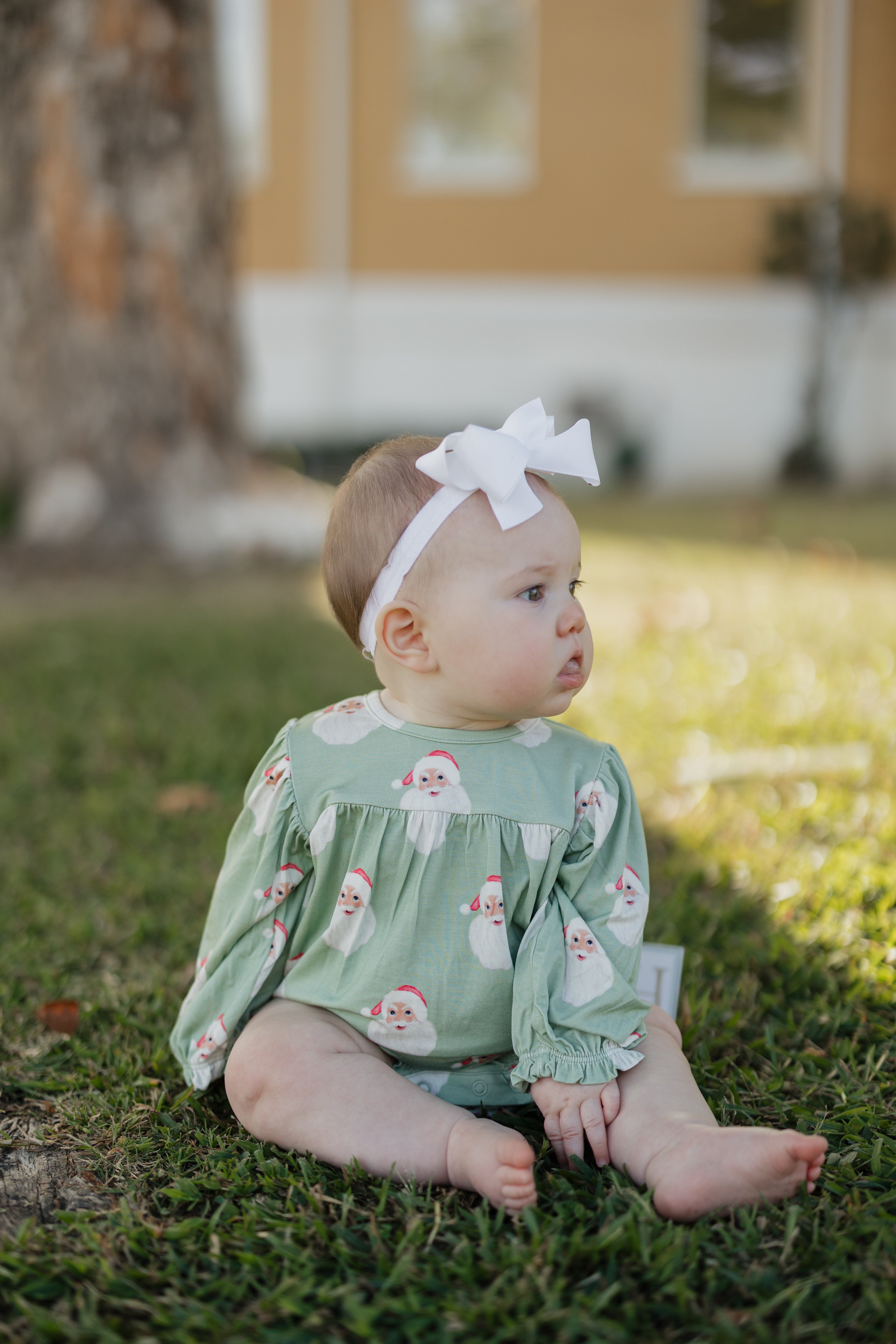 Baby sitting on grass wearing a green Santa bubble Romper with a white headband.