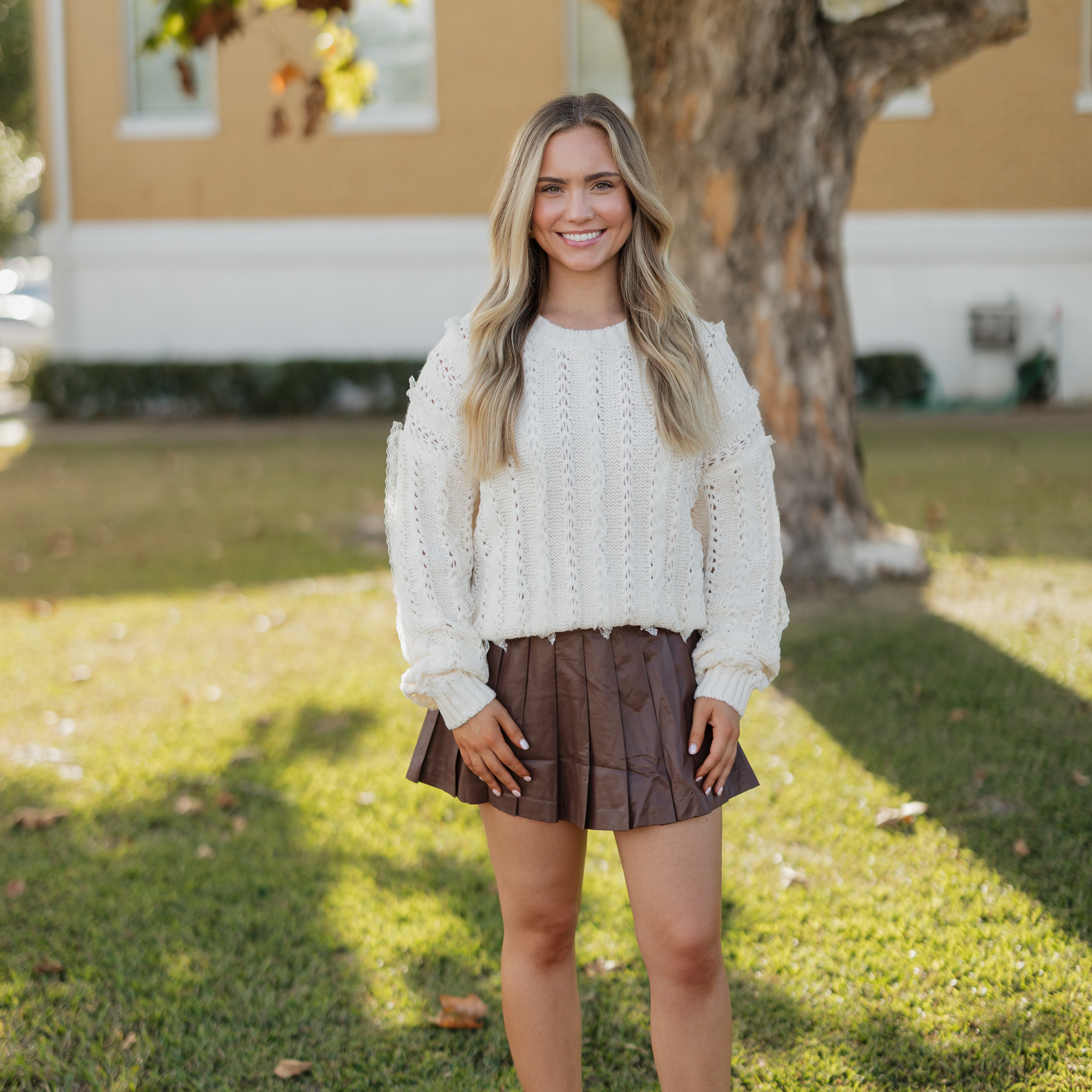 Woman in a white sweater and brown skirt standing outdoors with a tree and house in the background