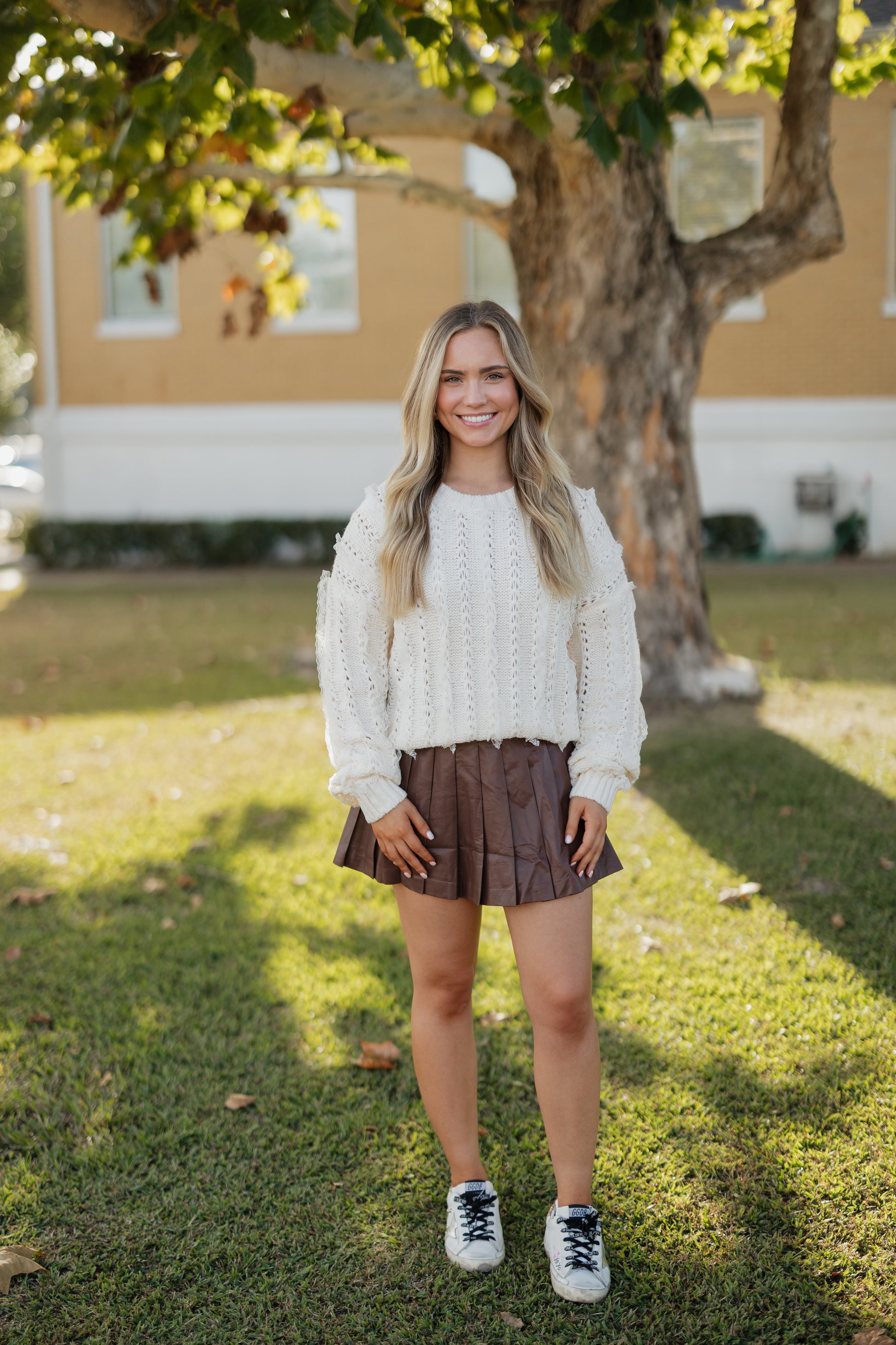 Woman in a white sweater and brown skirt standing outdoors with a tree and house in the background