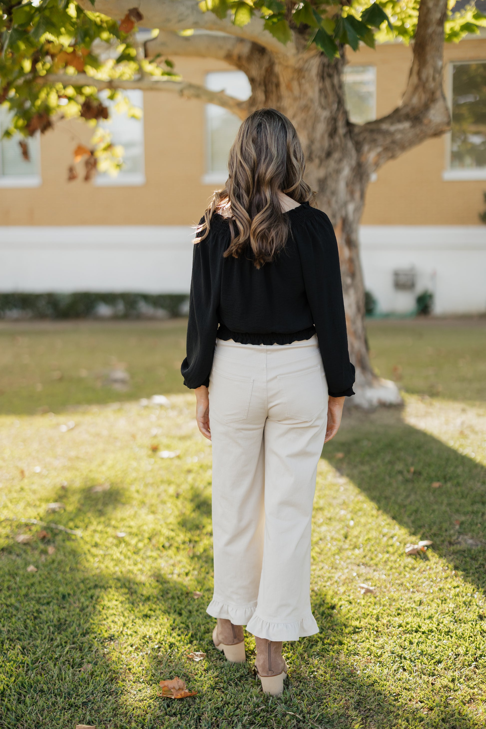 Person wearing a black top and white pants standing in a grassy area with a tree and building in the background.