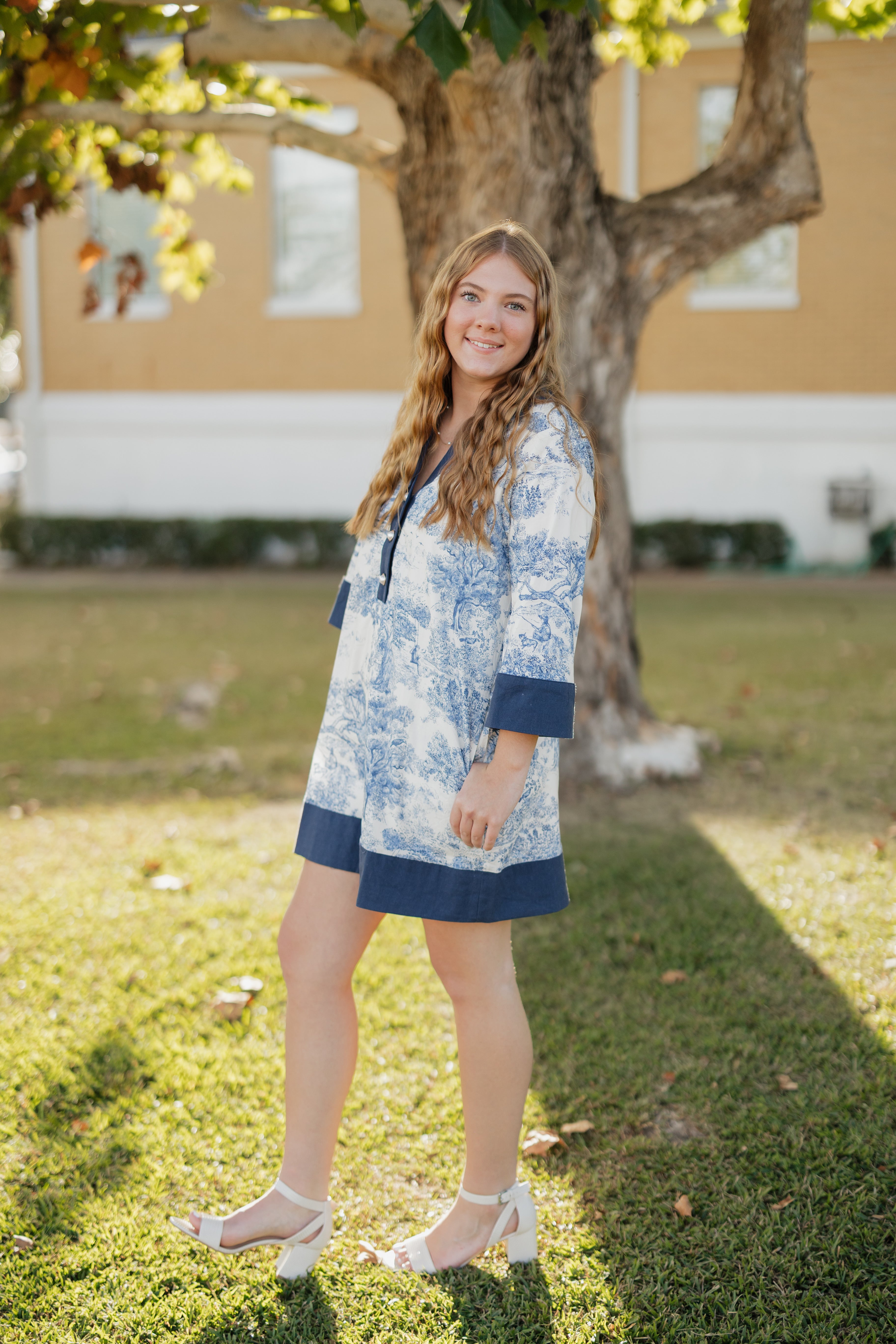 Woman in a blue and white floral dress standing outdoors near a tree