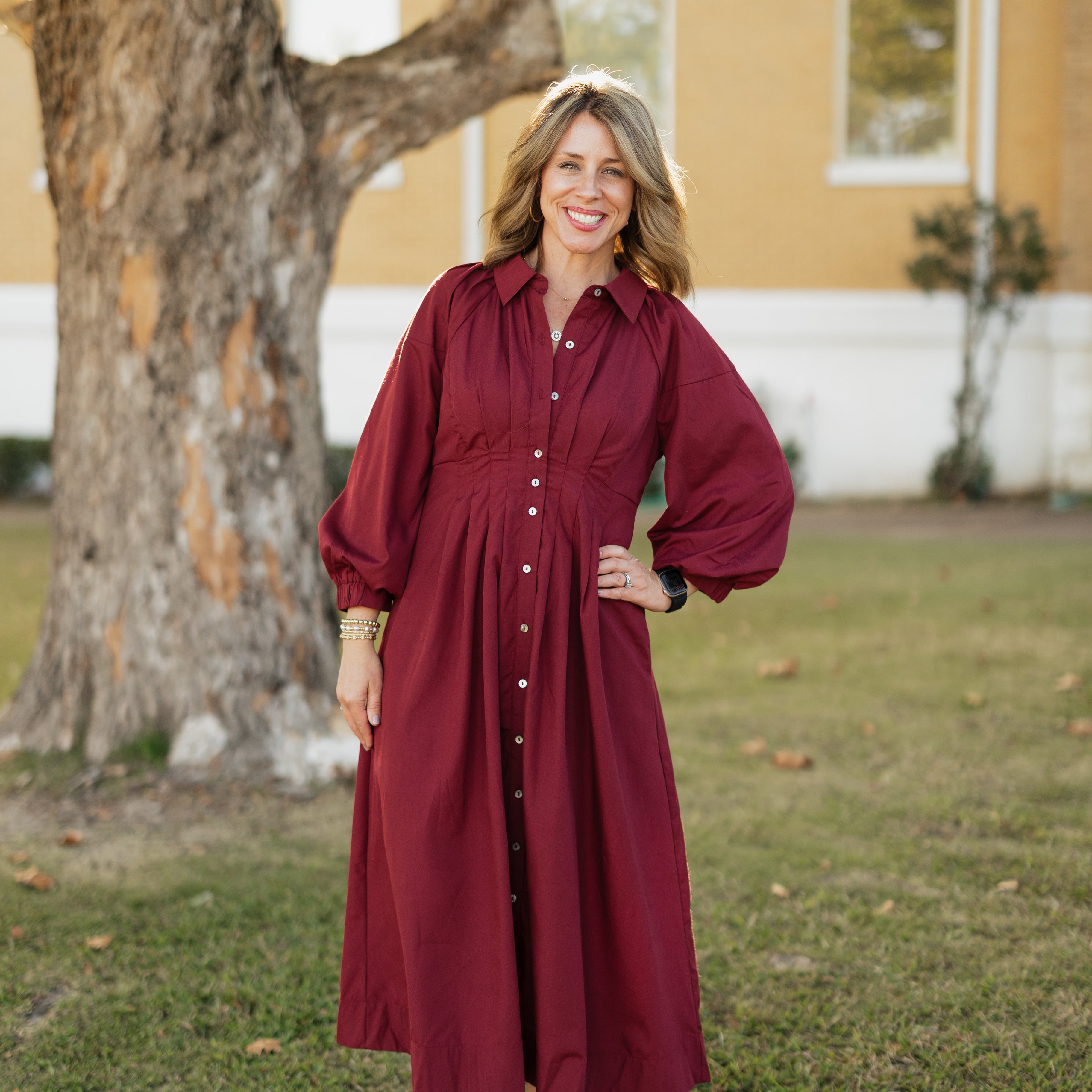 Woman in a garnet dress standing outdoors with a house and tree in the background