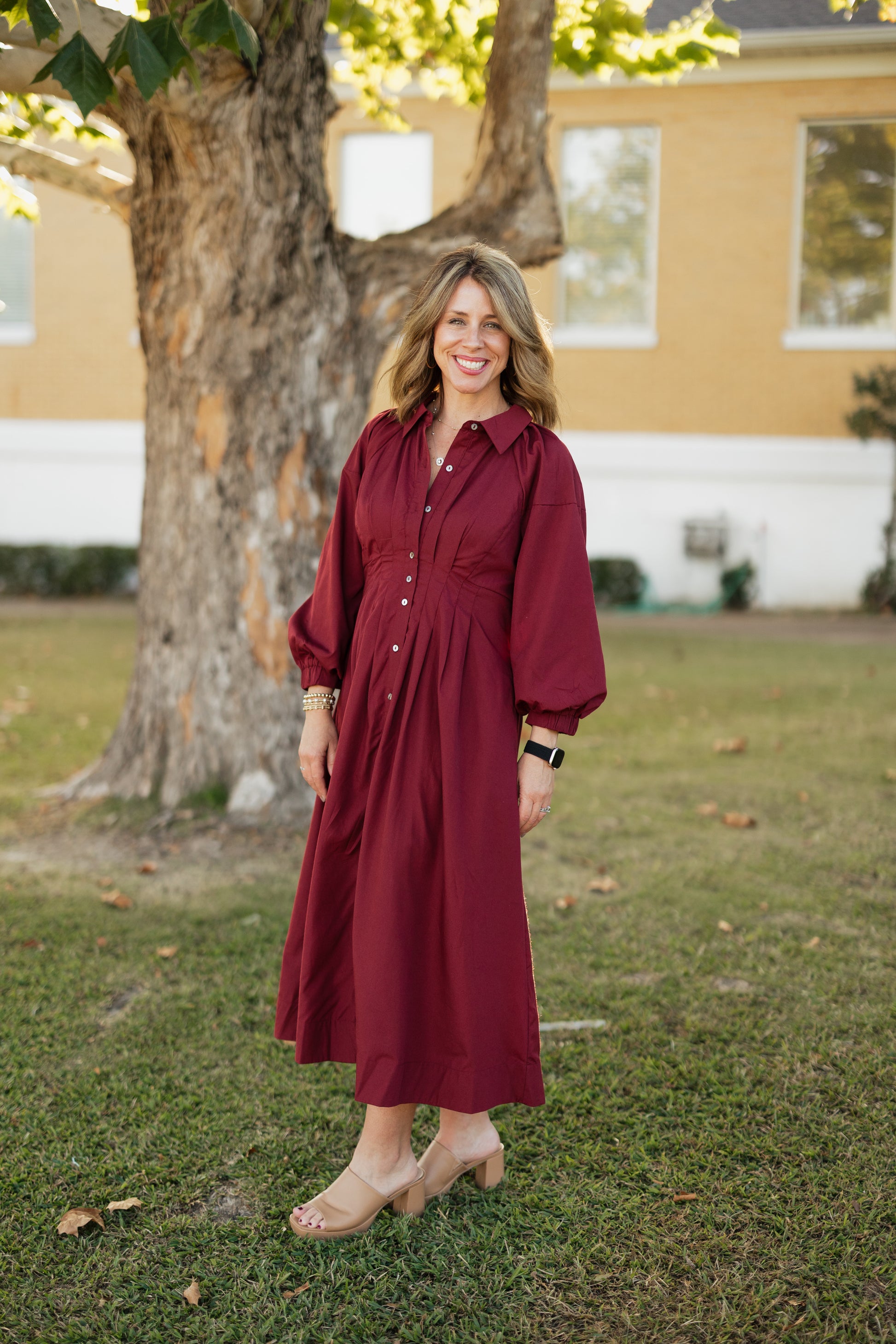 Woman in a garnet dress standing outdoors with a tree and house in the background