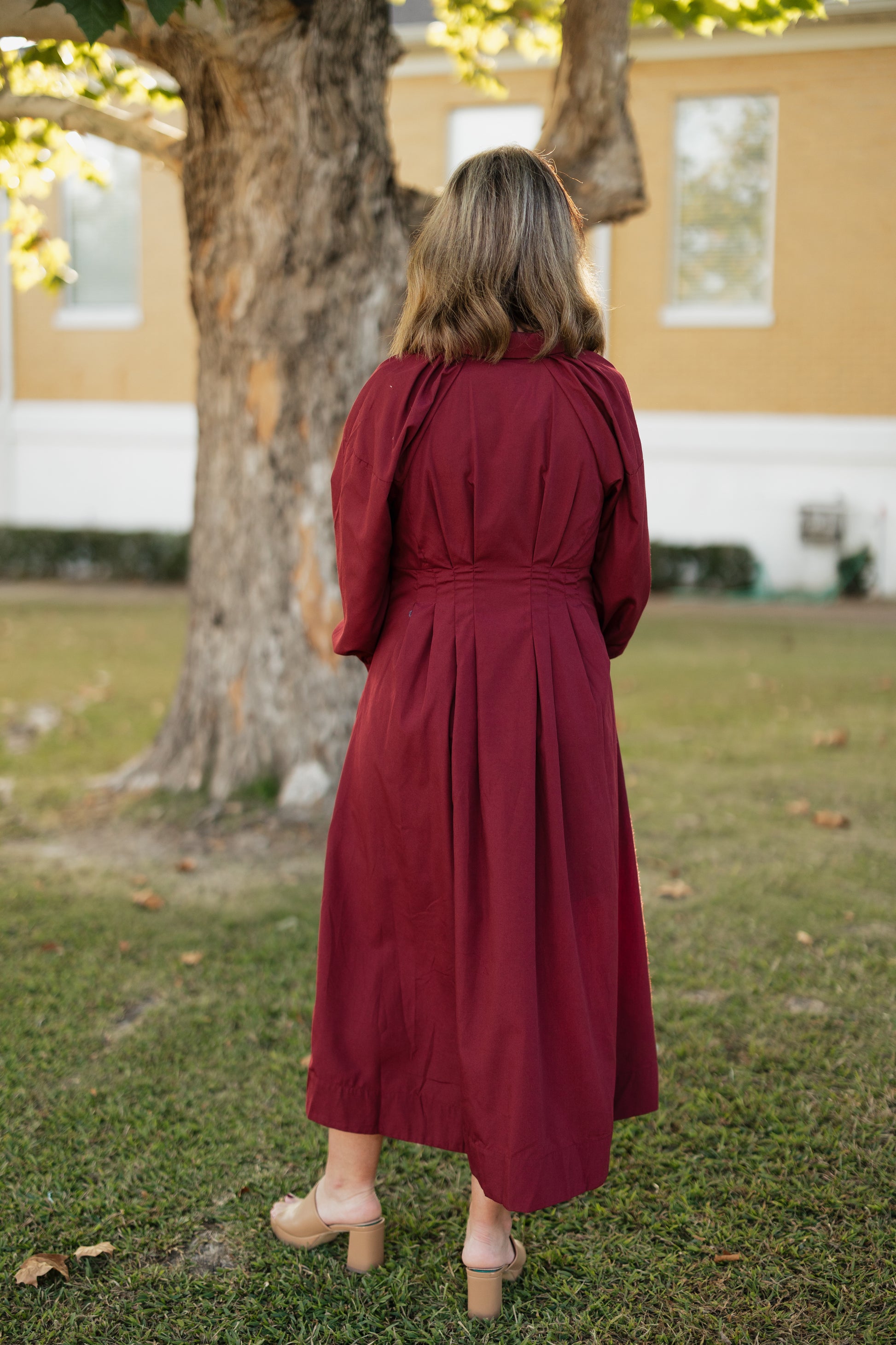 Woman in a garnet colored dress standing in a park-like setting with a tree and building in the background.