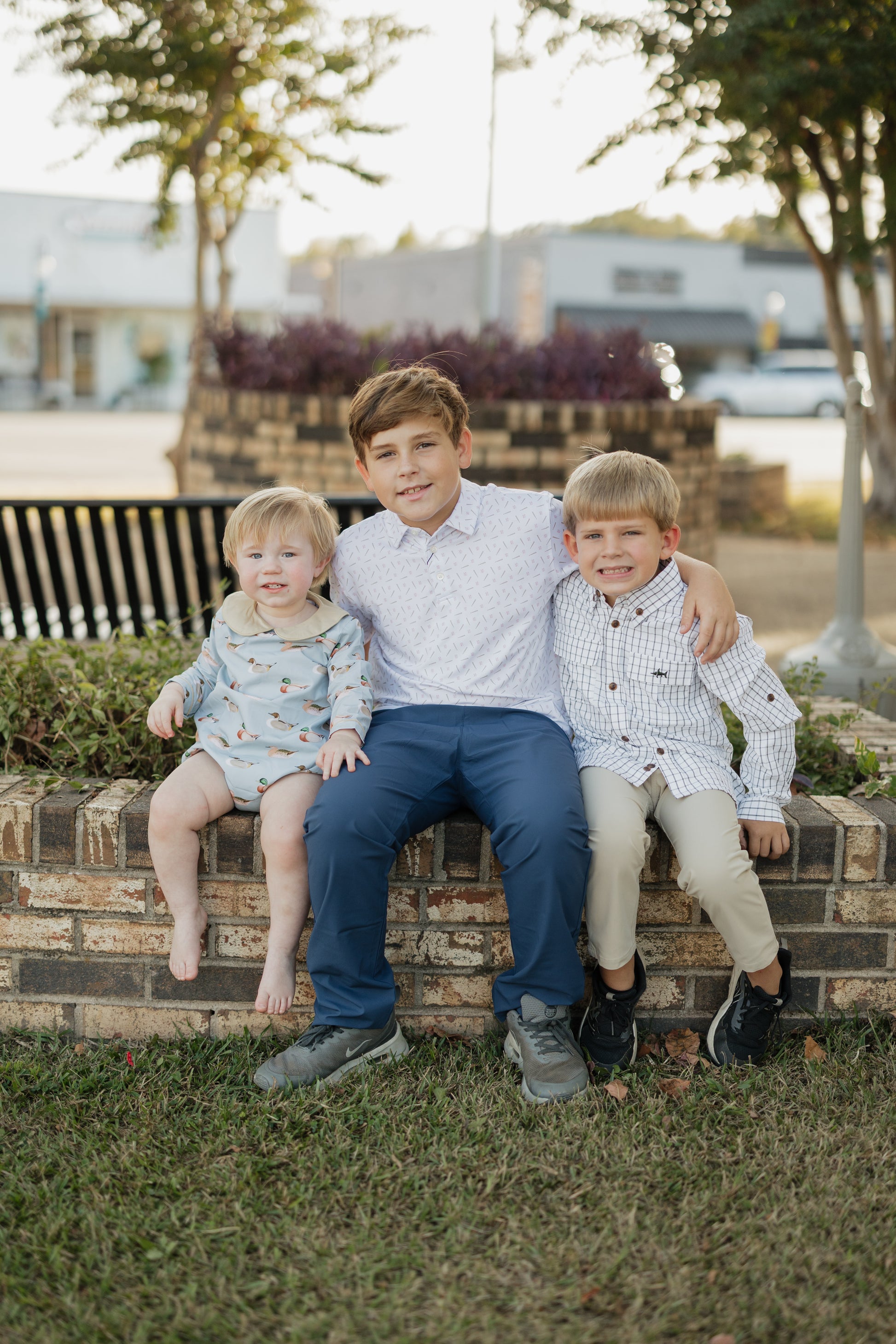 Three children sitting on a brick wall outdoors with trees and buildings in the background.