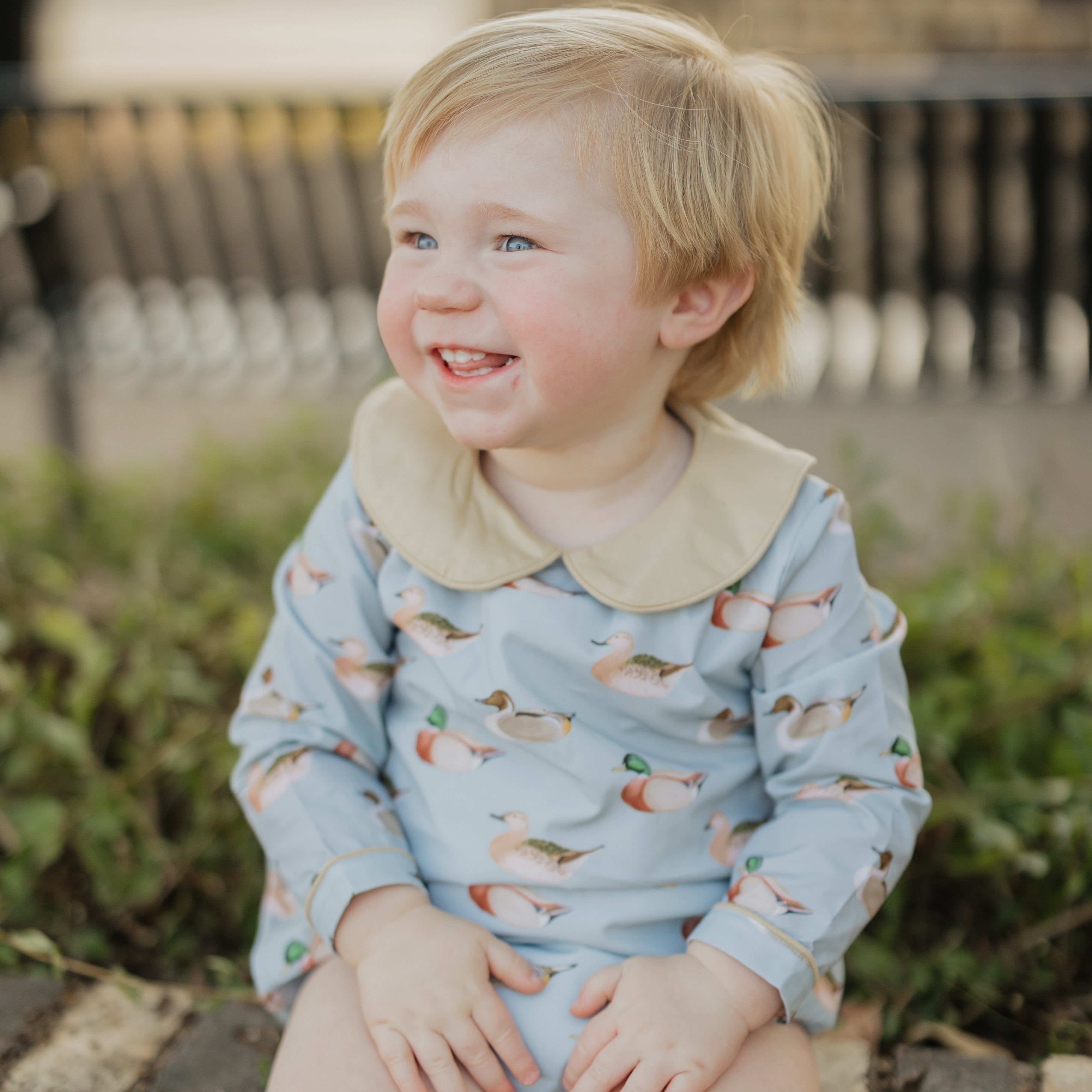 Child wearing a light blue onesie with animal patterns, sitting on a wooden bench outdoors.