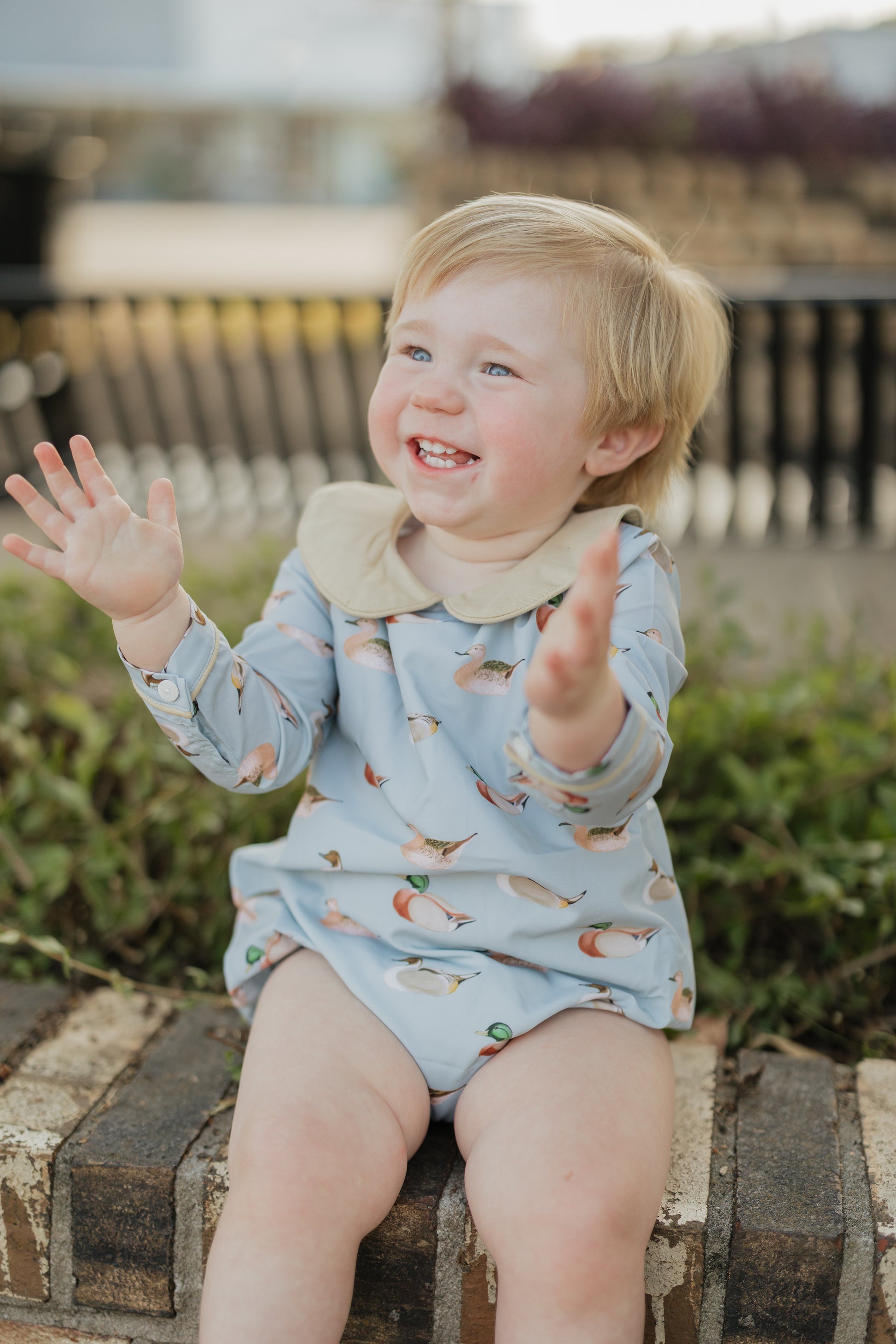 Child wearing a light blue romper with animal prints, sitting on a wooden bench outdoors.